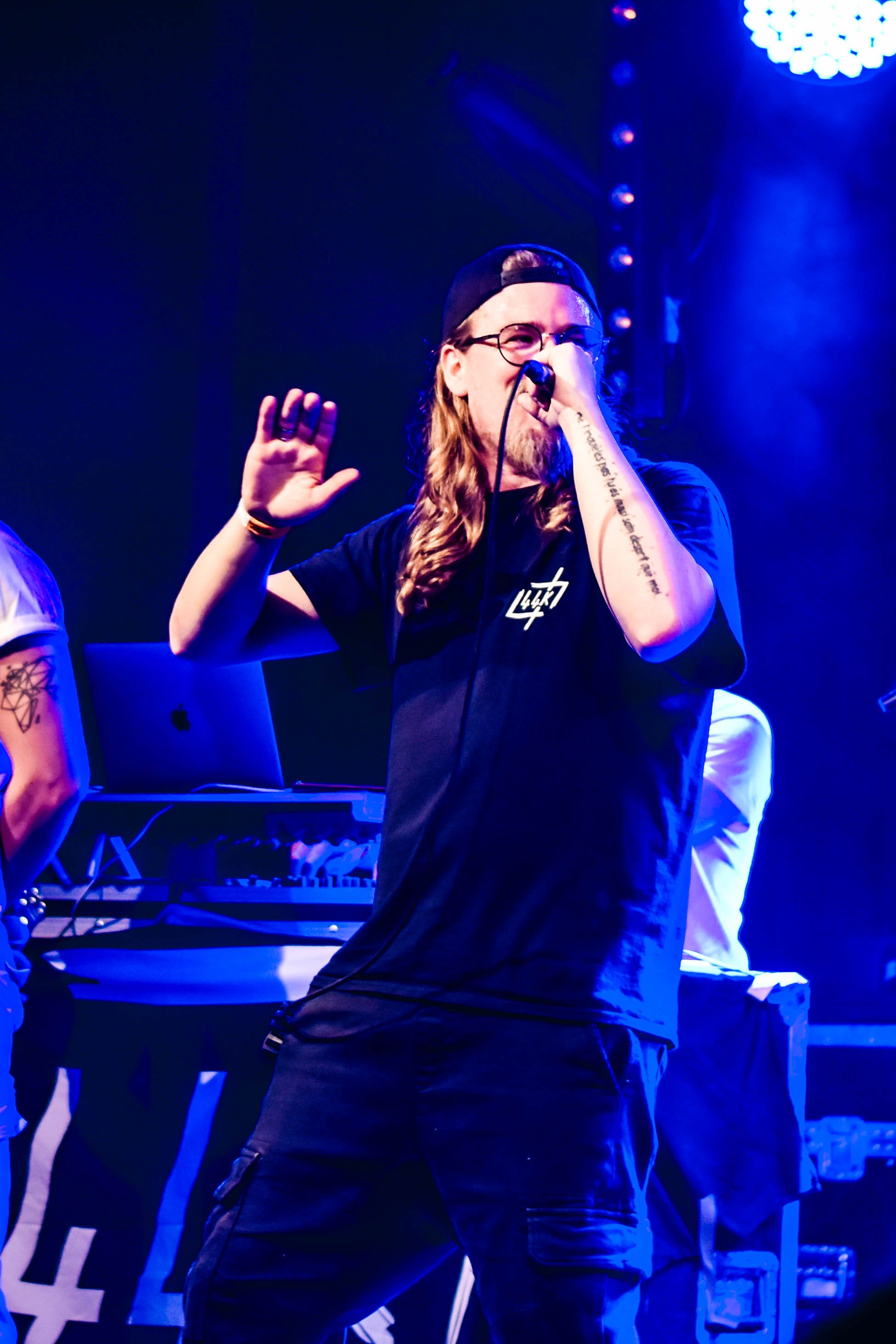 Un homme avec des longs cheveux, lunettes, portant un t-shirt noir avec un logo blanc, tenant un microphone lors d'une performance musicale sur scène avec des lumières bleues. 
