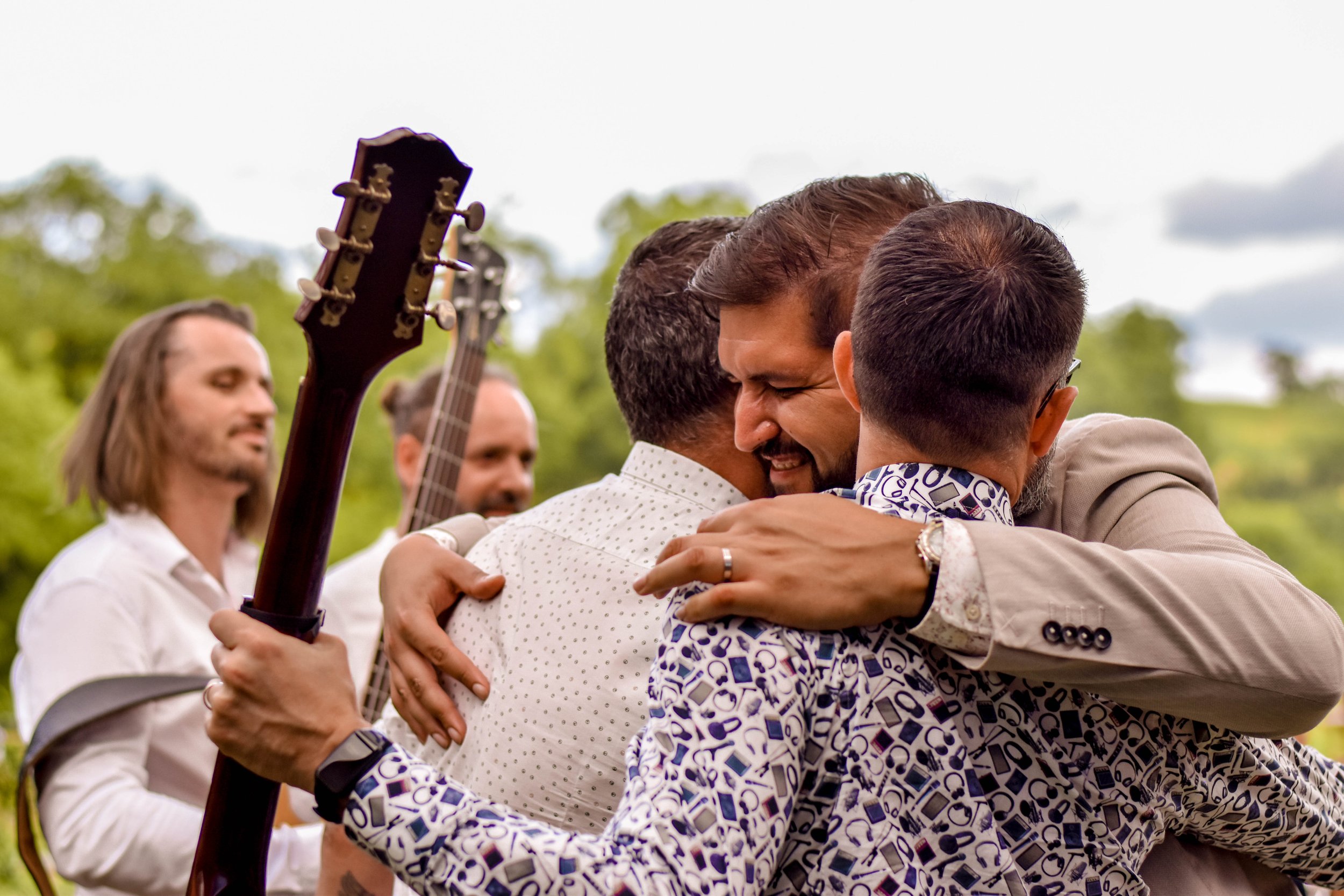 Trois hommes qui se prennent dans les bras, avec un groupe de musiciens en arrière-plan, dans un cadre extérieur naturel.