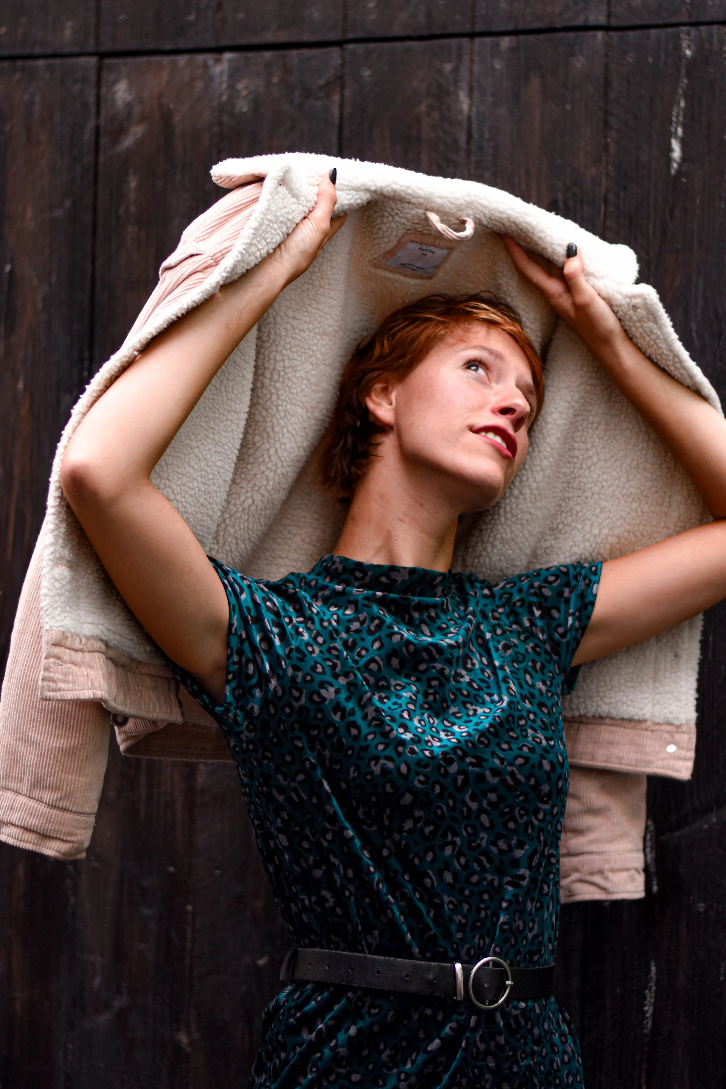 Une femme aux cheveux roux porte une robe à motifs léopard bleus et noirs, un ceinture noire avec boucle métallique, et est en train de retirer ou d'enrouler une veste ou un manteau en peluche blanc, le tout contre un fond de bois sombre.