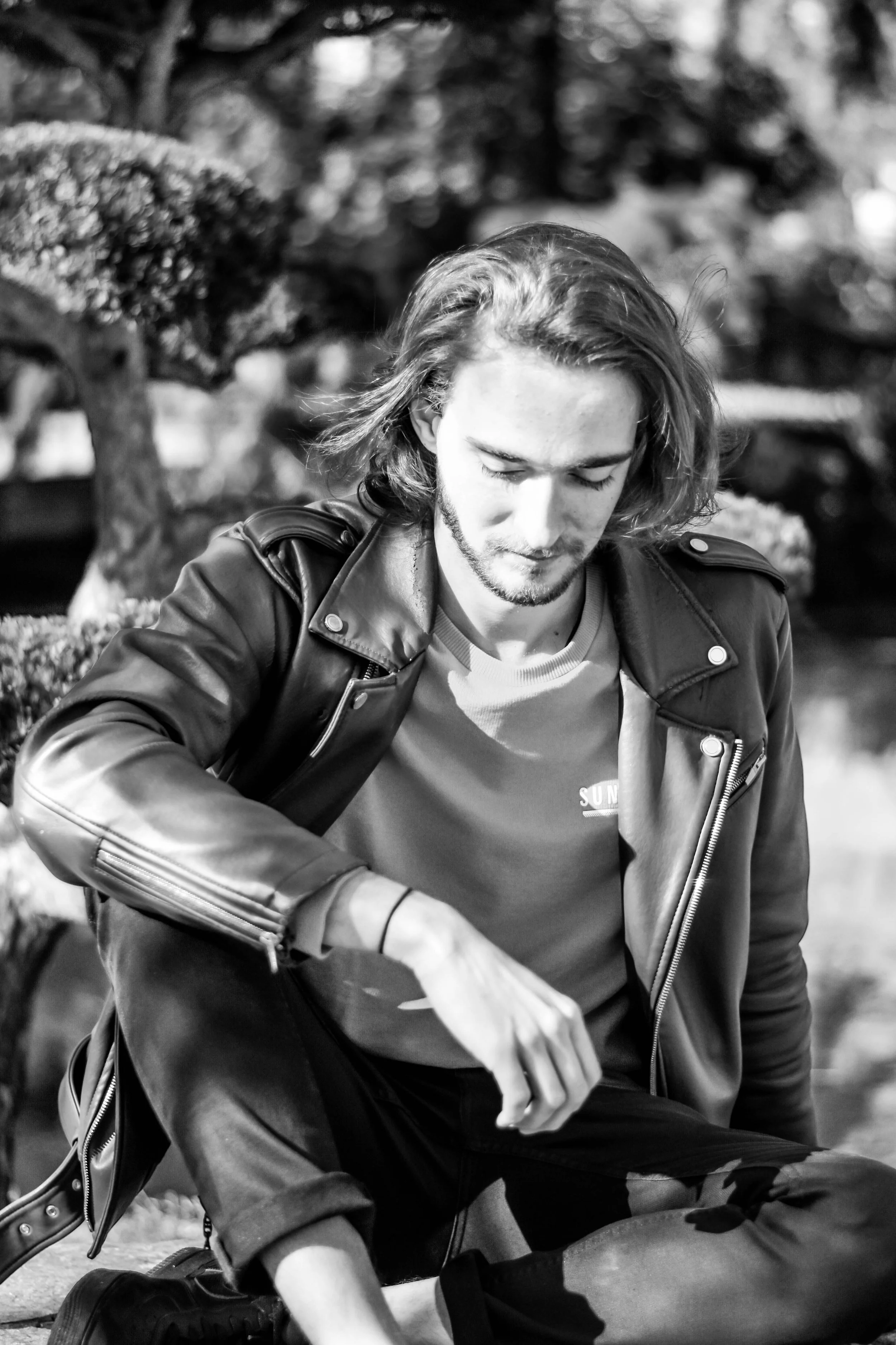 Un jeune homme aux cheveux longs, portant une veste en cuir et un t-shirt, est assis avec les yeux fermés, dans un environnement naturel, probablement dans un parc.