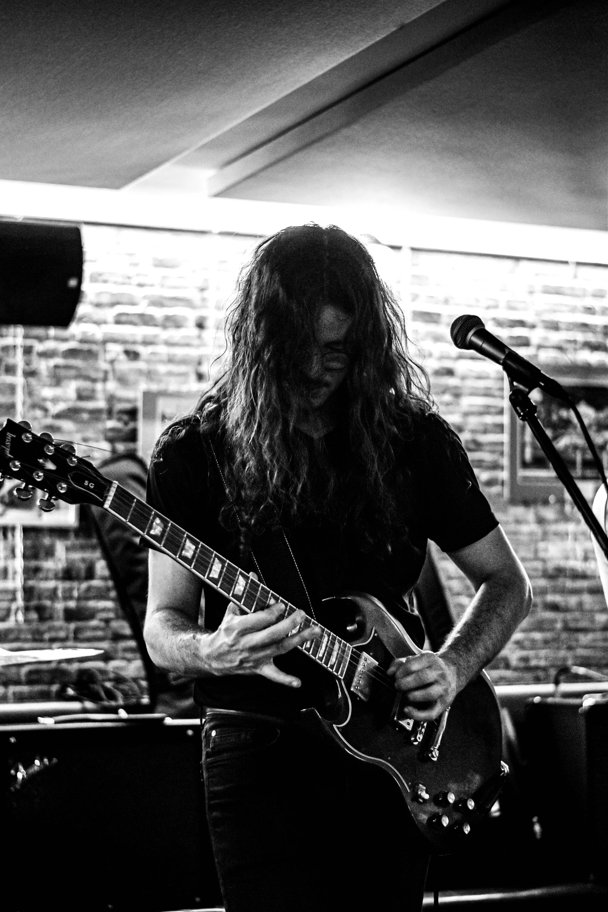 Musicien jouant de la guitare électrique dans un local de musique avec un mur en briques en arrière-plan, en noir et blanc.