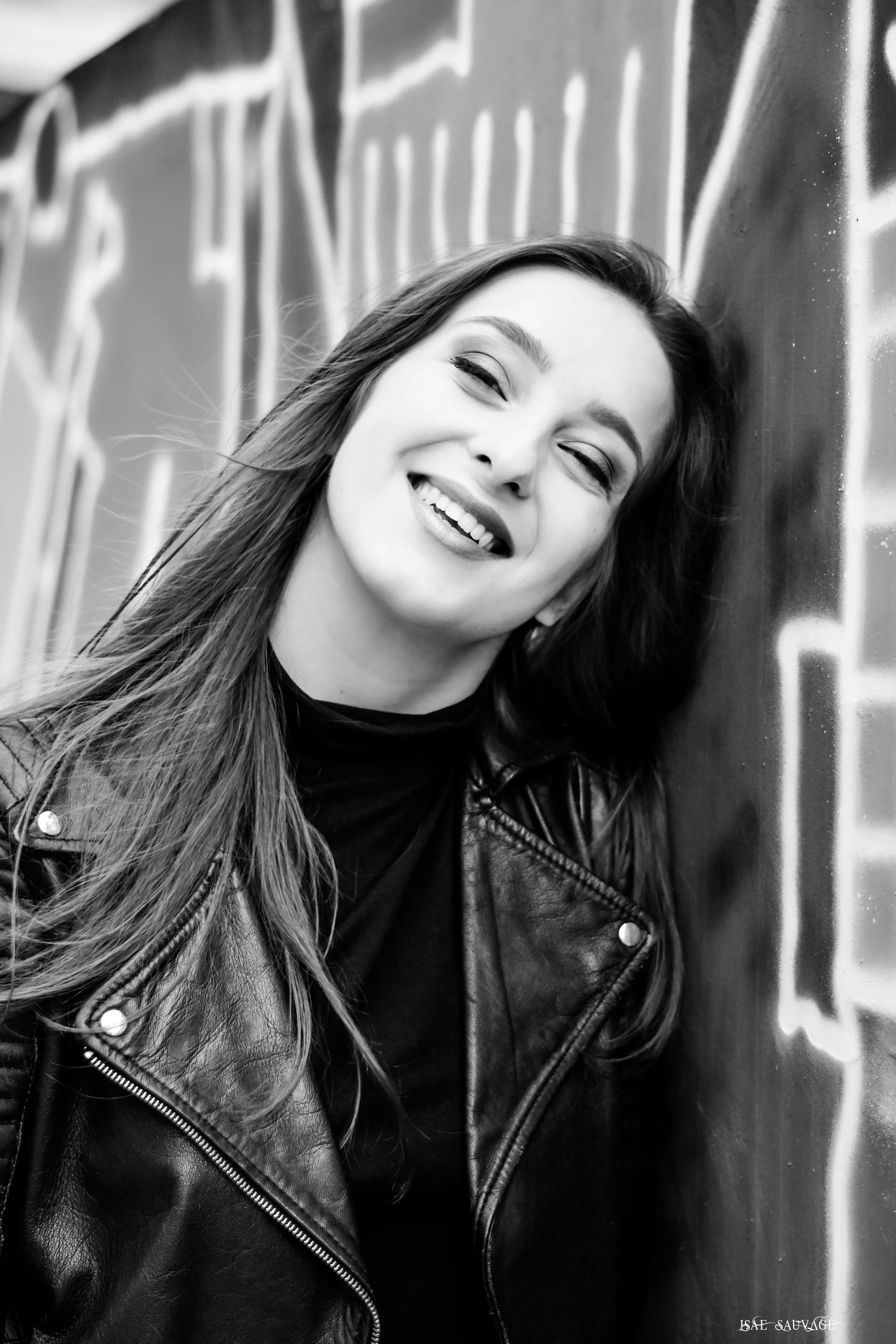 Une femme souriante portant une veste en cuir noir, posant contre un mur de graffiti