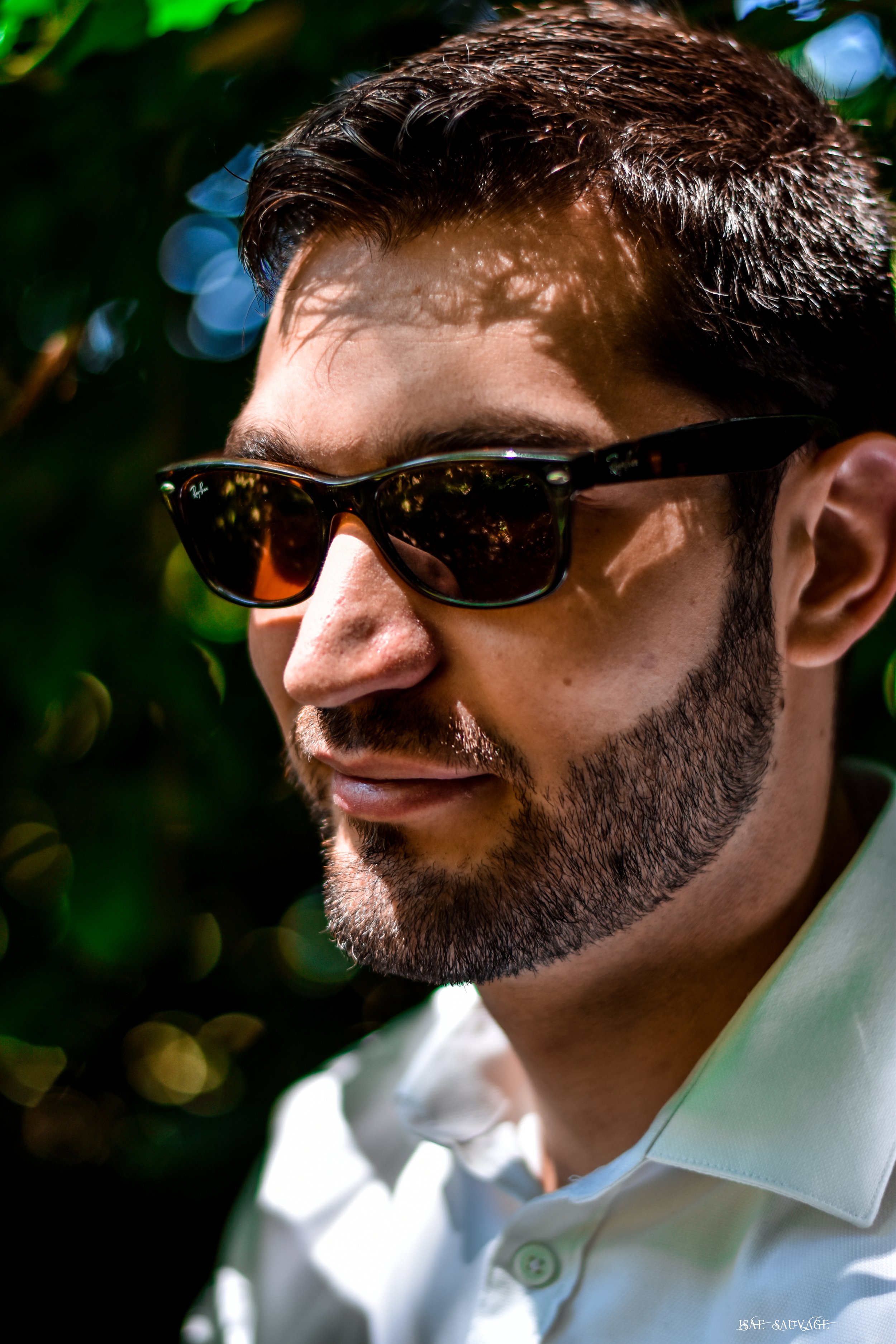 Jeune homme portant des lunettes de soleil et une chemise blanche, avec des cheveux courts et une barbe, sous un arbre.