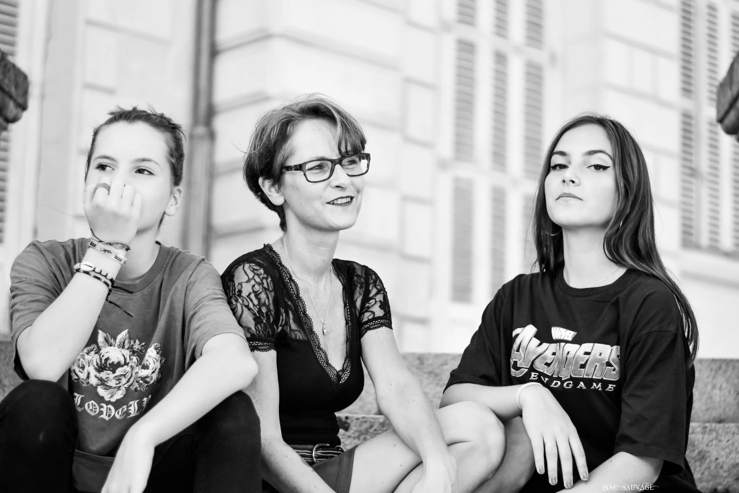 Trois jeunes femmes assises côte à côte dans un environnement urbain. La femme au centre porte des lunettes et un haut noir avec des manches en dentelle. La femme à gauche porte un t-shirt avec une fleur imprimée et plusieurs bracelets, elle couvre s