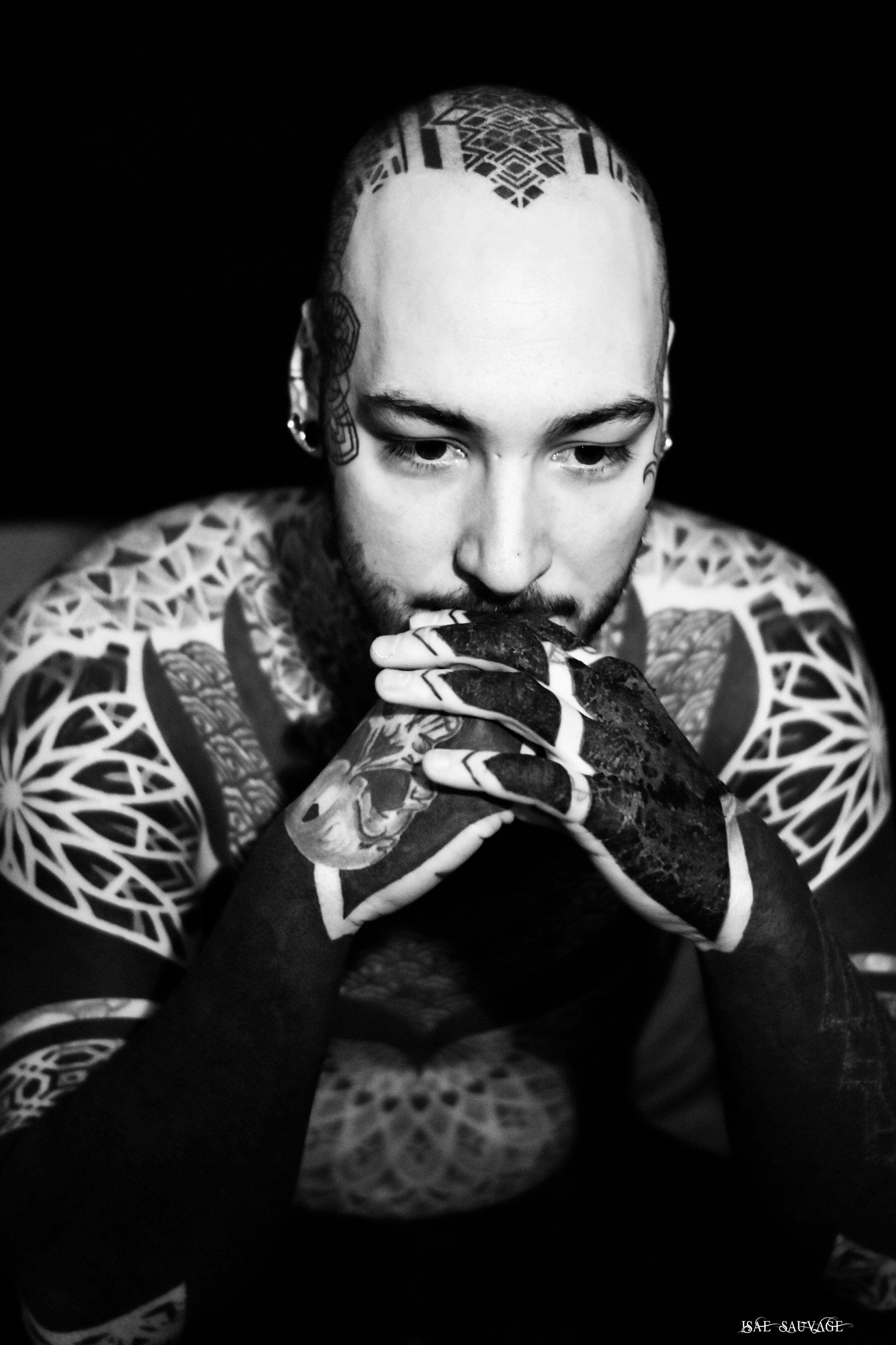 Un homme tatoué avec la tête rasée, regardant vers le bas, tenant sa main près de la bouche dans une pose contemplative, en noir et blanc.