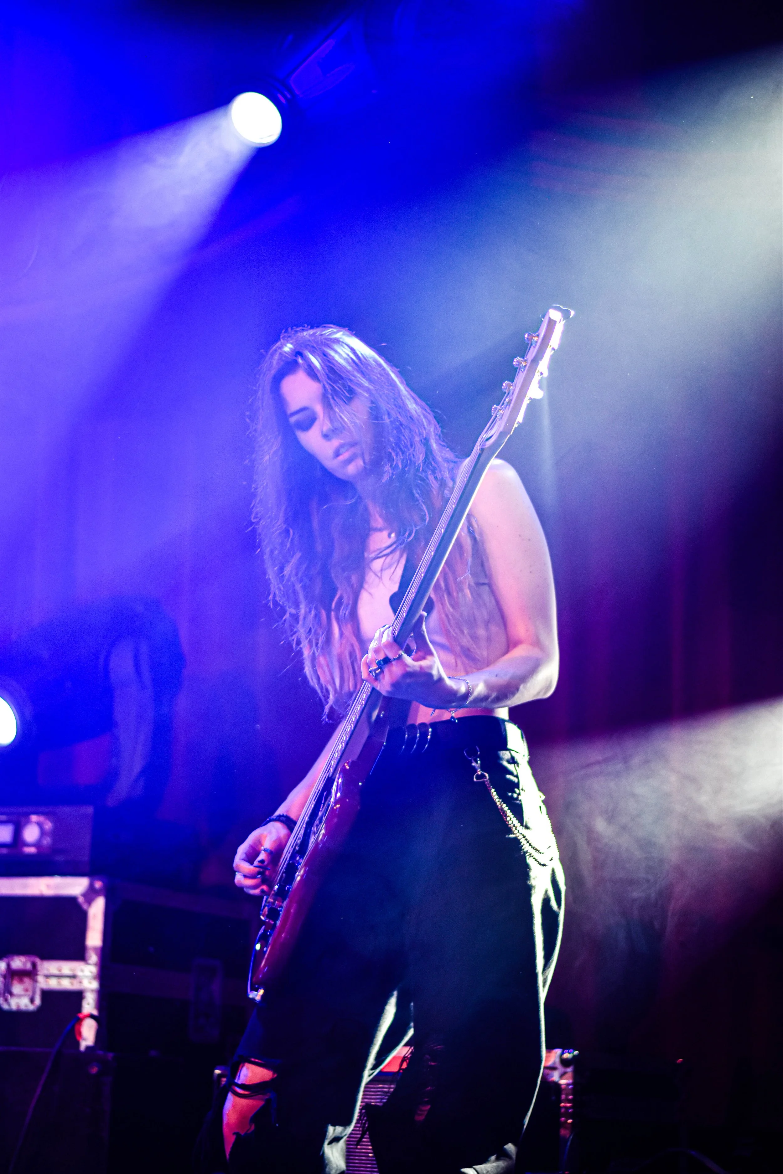 Une femme avec des cheveux longs et bouclés joue de la guitare électrique sur scène, sous des lumières violettes et blanches, lors d'une performance musicale.