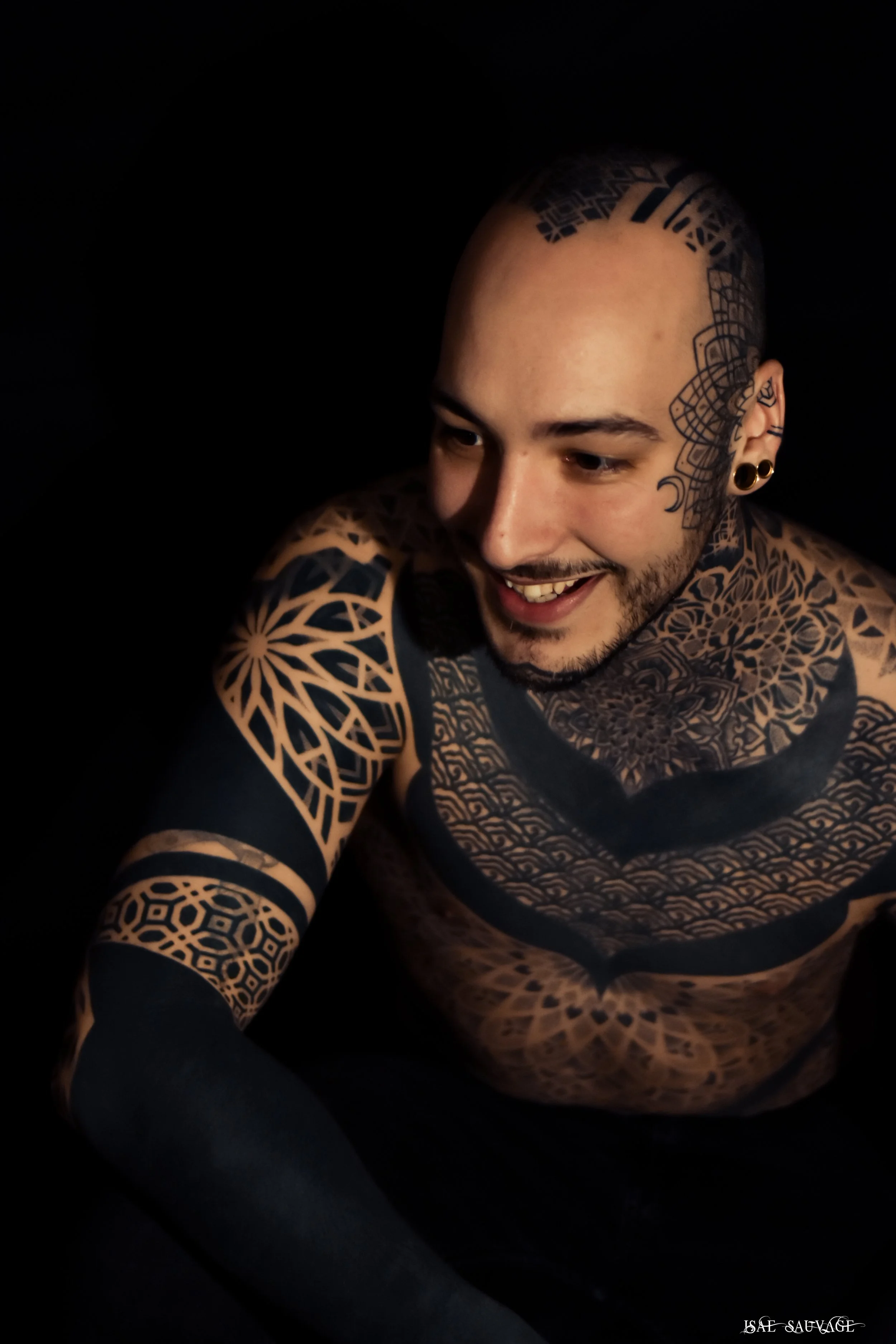 Un homme souriant avec de nombreux tatouages, une tête rasée, portant des boucles d'oreilles à cercles noirs, pose dans l'obscurité.