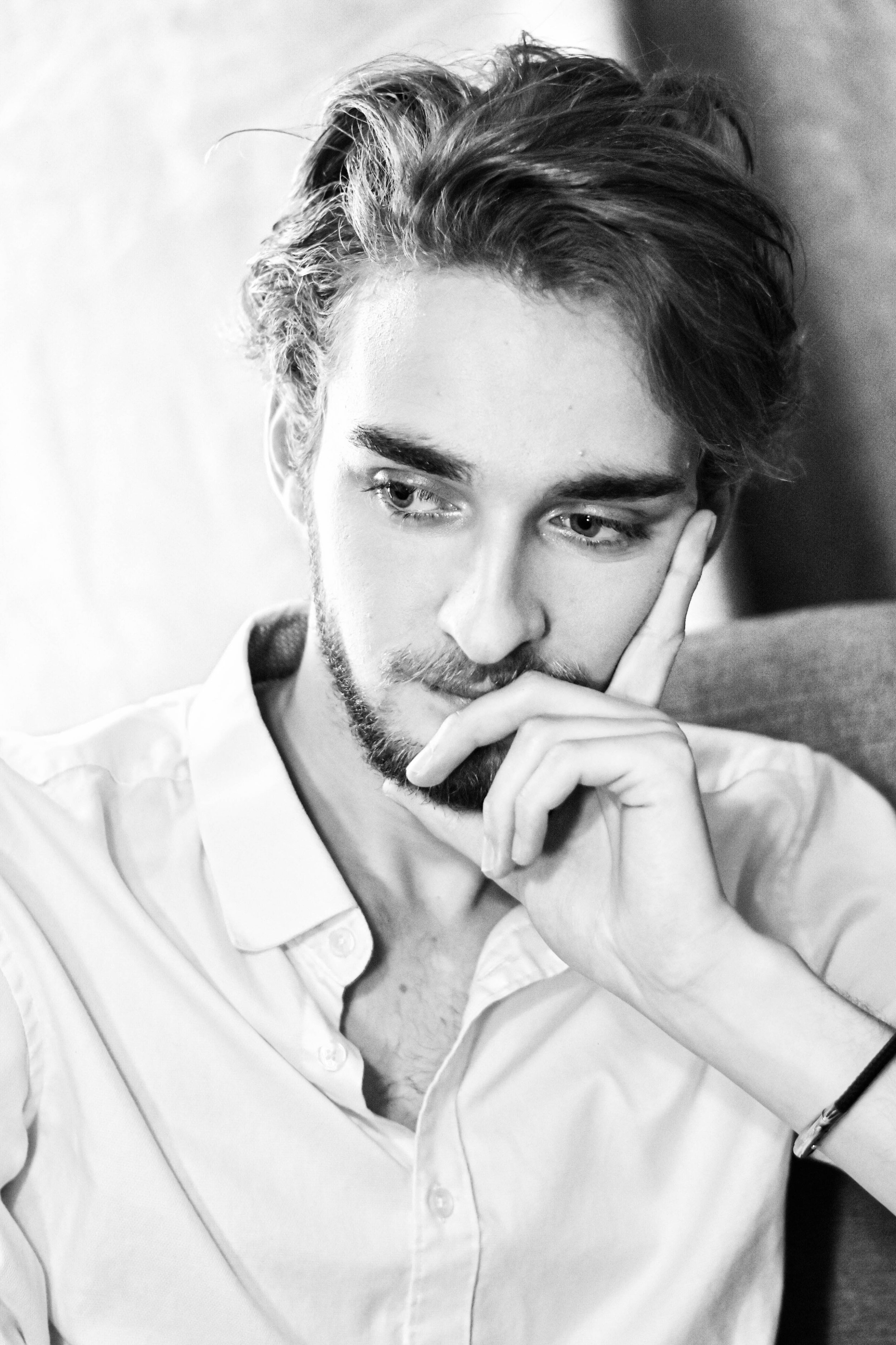Un jeune homme aux cheveux coiffés, portant une chemise blanche, en train de penser, avec la main près du visage.
