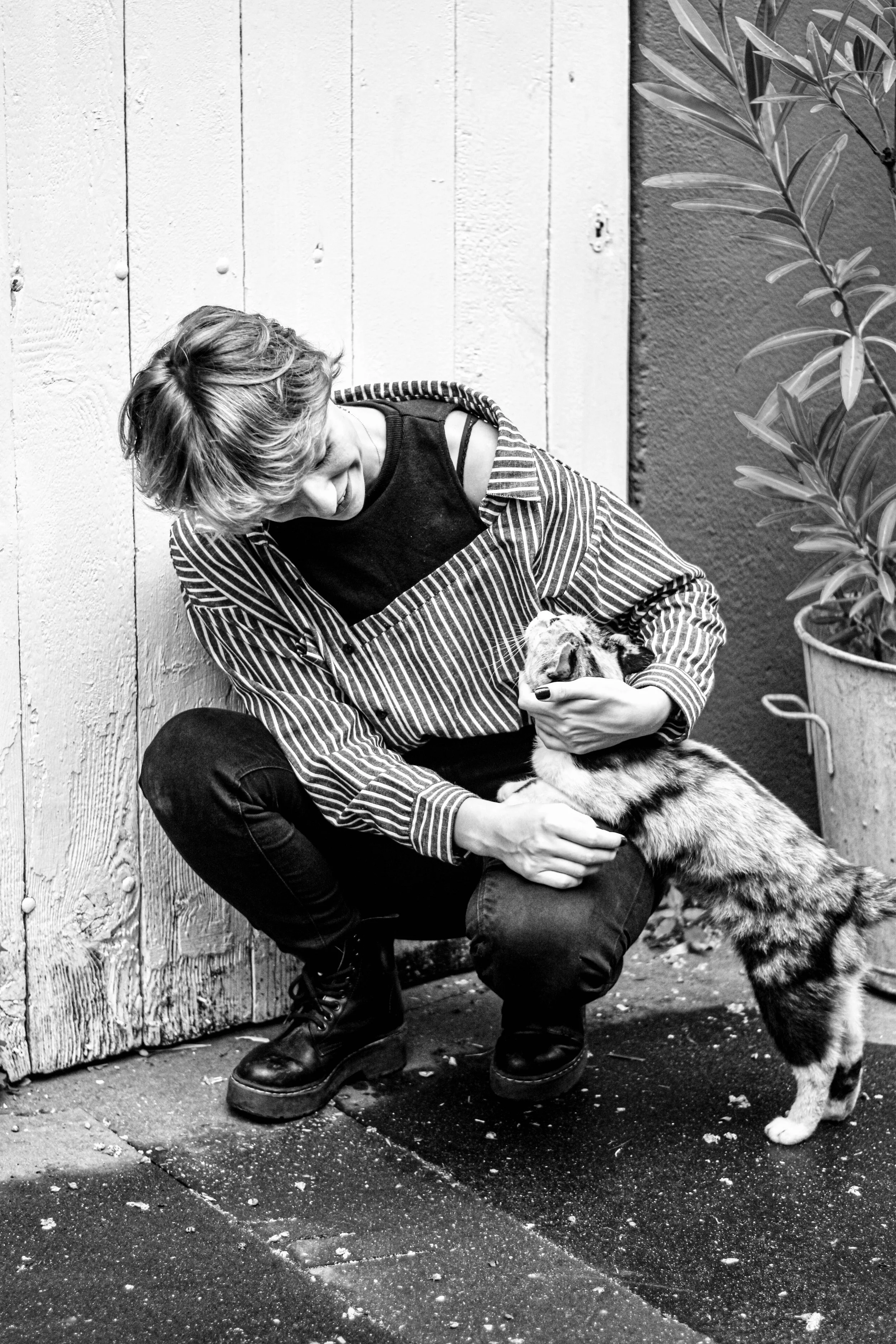 Une femme assise près d'une porte en bois, caressant un chien dans la rue. La scène est en noir et blanc, avec une plante à droite et un sol en béton.