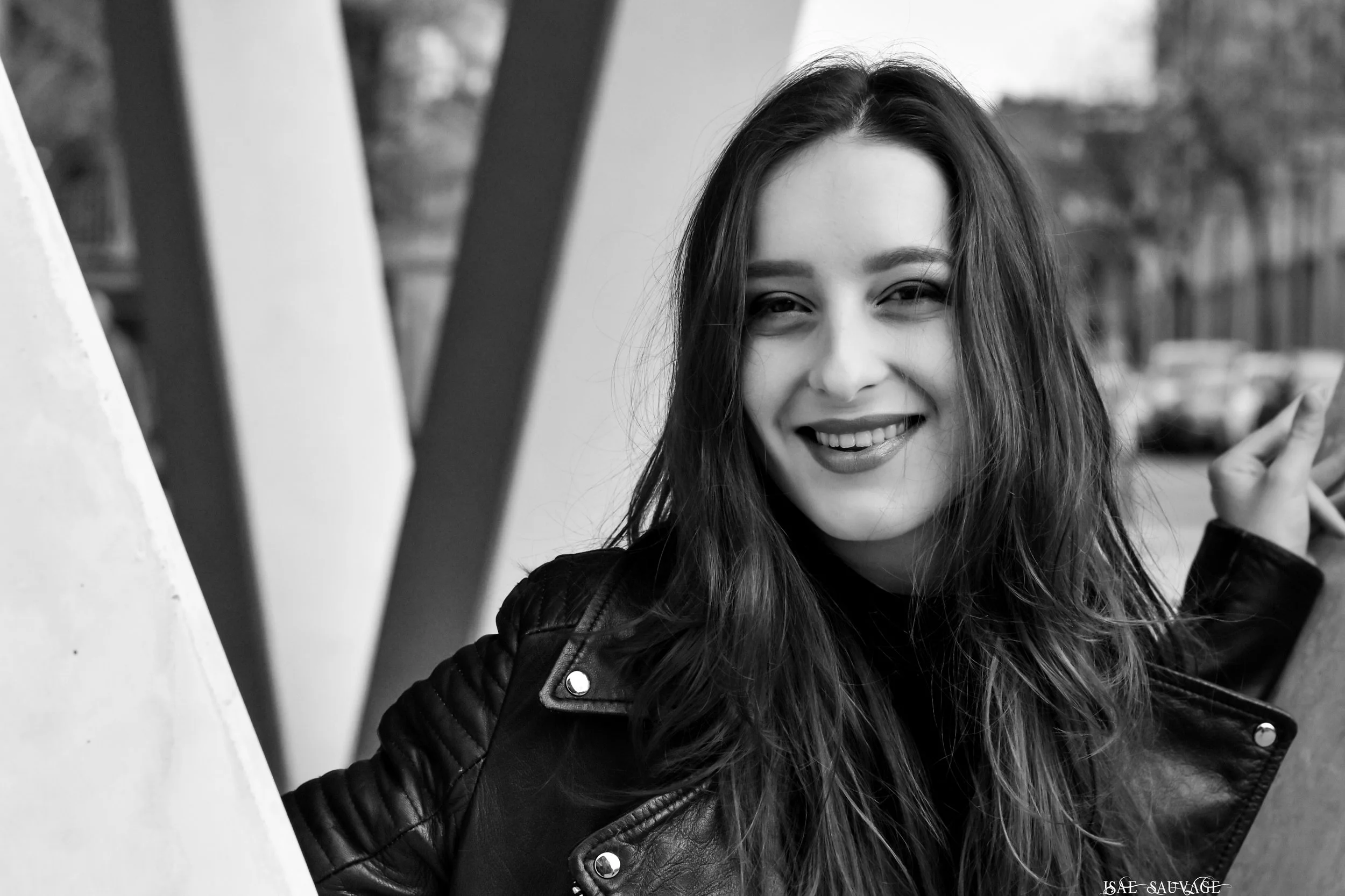 Jeune femme souriante avec cheveux longs et vêtements en cuir, prise en photo en noir et blanc lors d'une journée en extérieur.