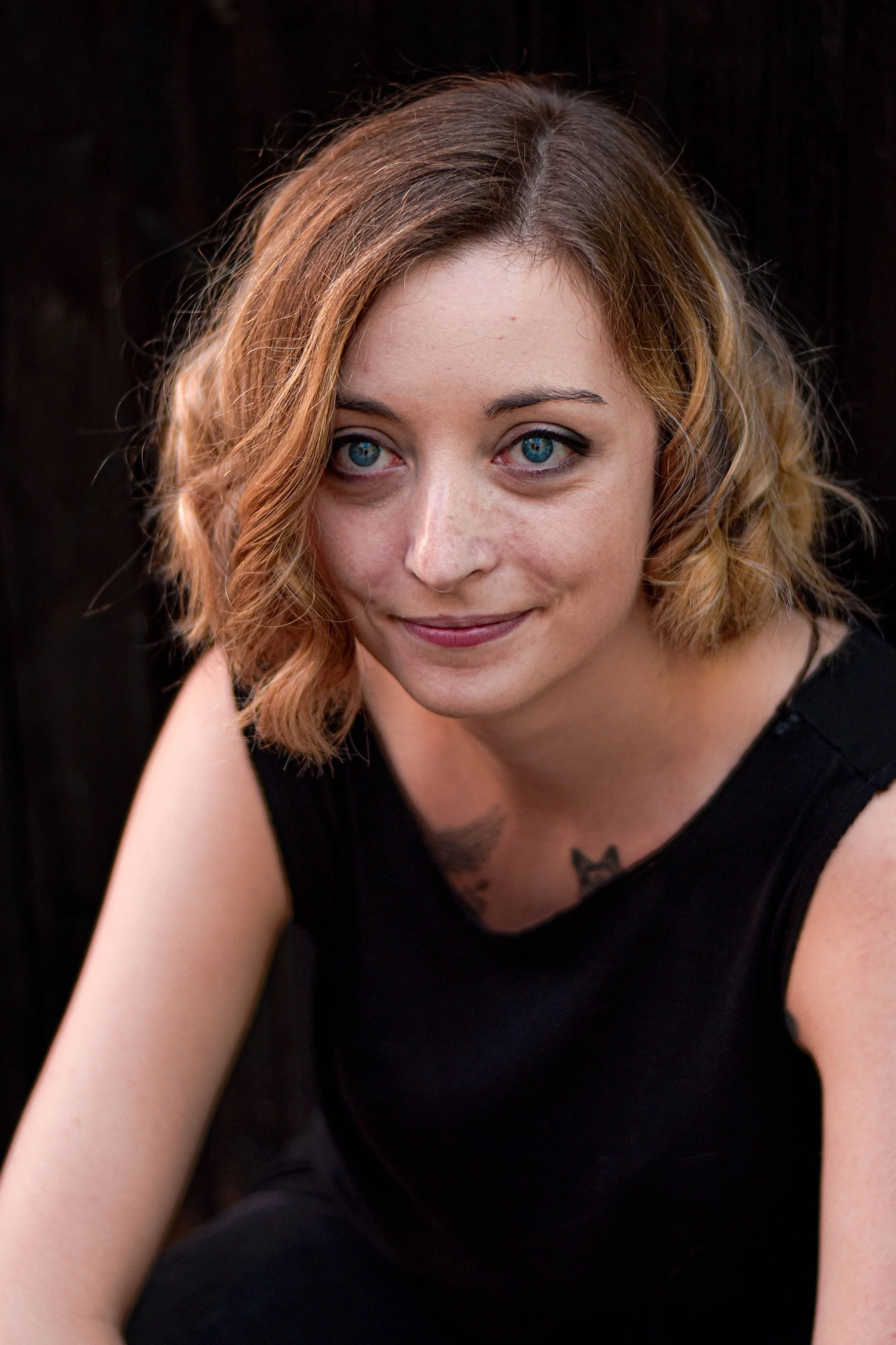 Jeune femme aux cheveux bruns bouclés et aux yeux bleus, portant un haut noir, assise contre un fond sombre, regardant l'objectif avec un léger sourire.