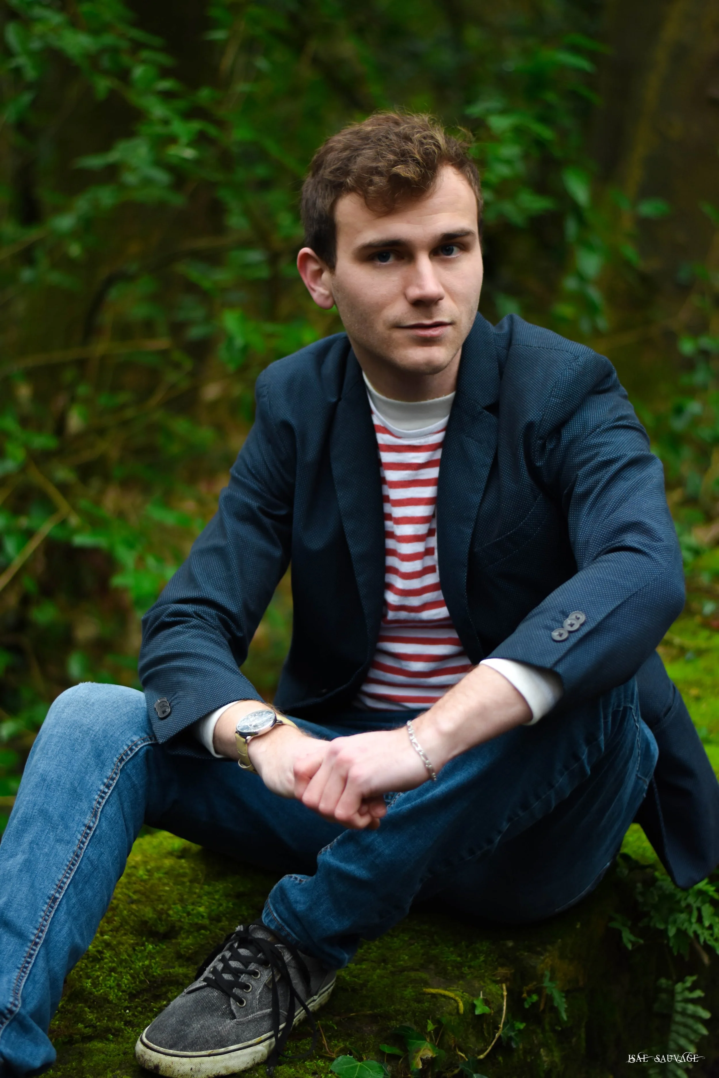 Un jeune homme assis sur la nature, portant une veste bleue, un T-shirt à rayures et des baskets, dans un environnement forestier vert.