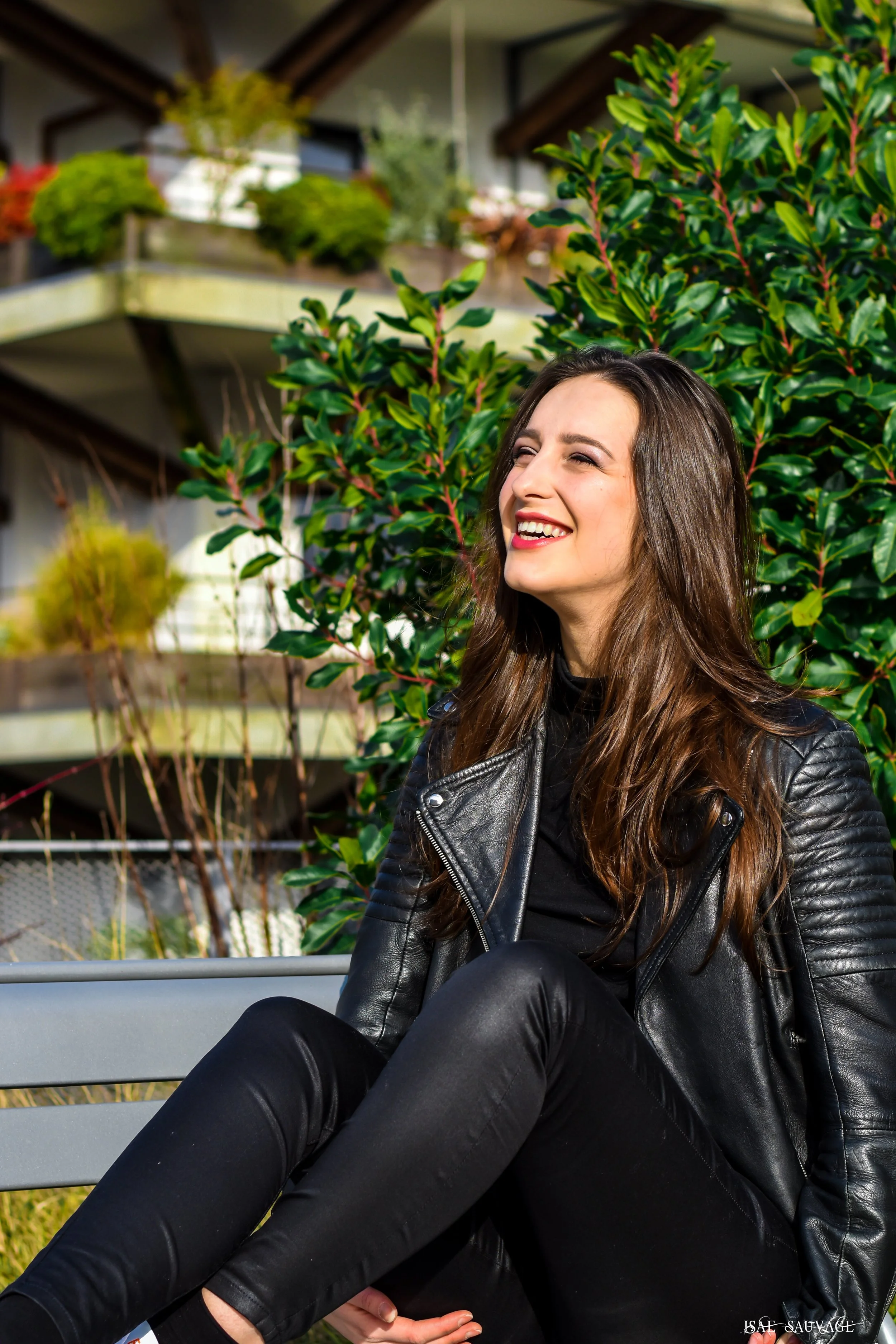 Une femme souriante avec des cheveux bruns, portant une veste en cuir noir, assise en extérieur devant des plantes vertes.