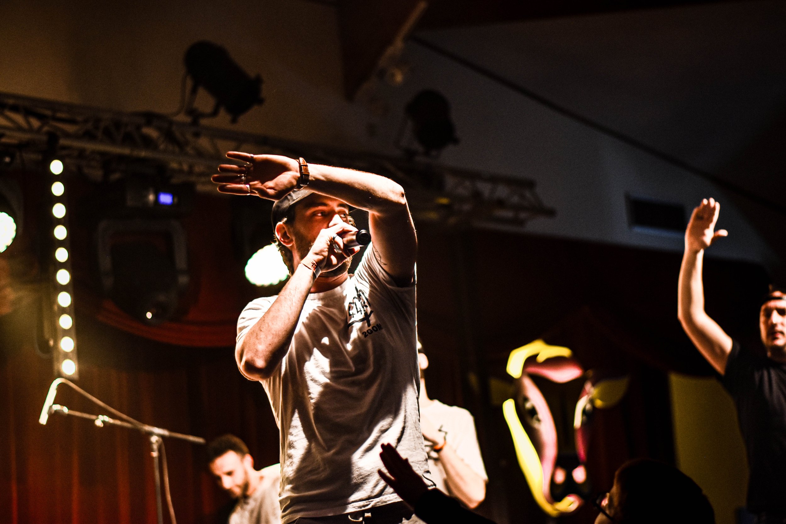 Un chanteur avec un micro sur scène, portant un t-shirt gris, lors d'un concert, avec d'autres personnes en arrière-plan.