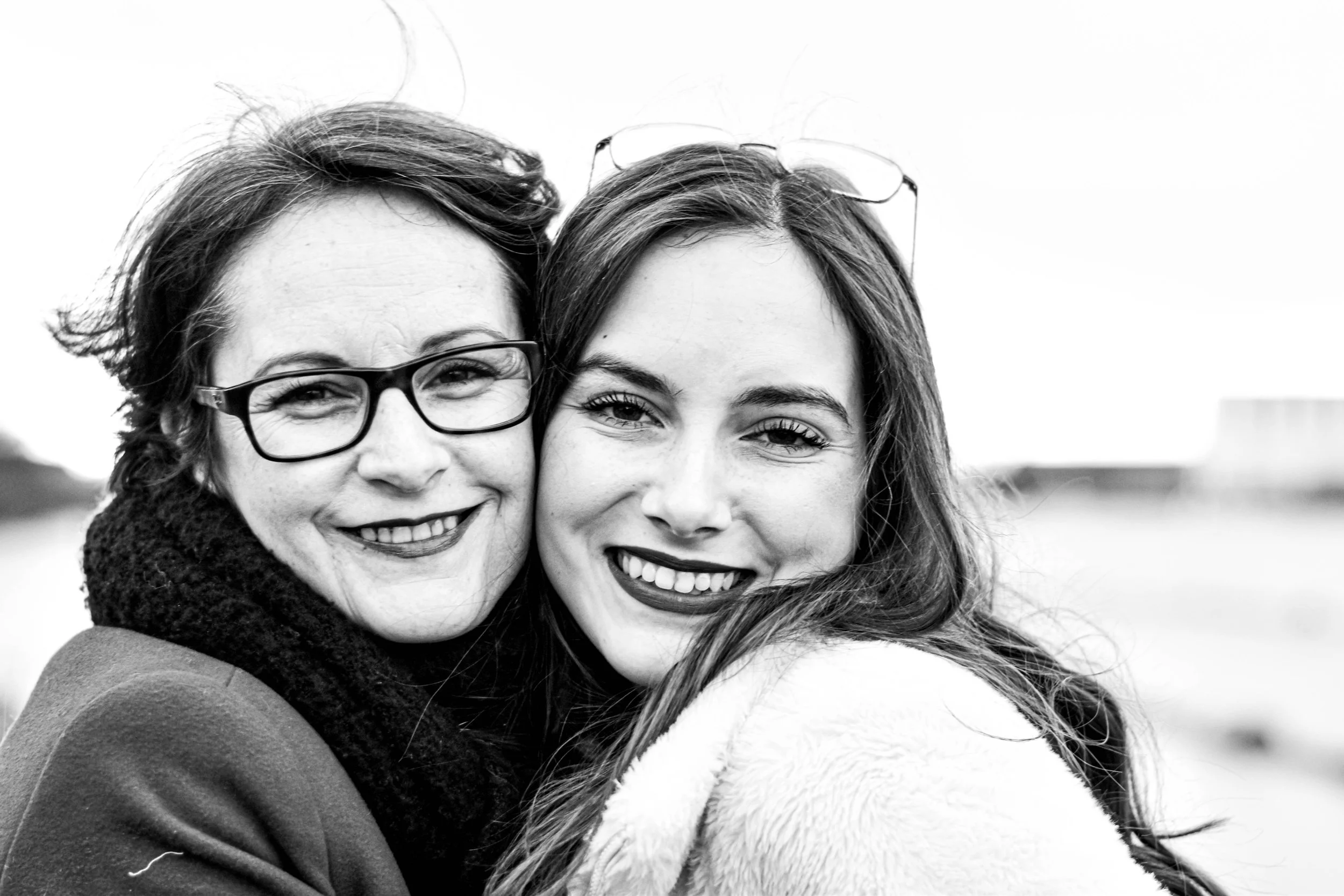 Une mère et sa fille, toutes deux souriantes, une porte des lunettes et l'autre ne porte pas de lunettes, en gros plan, en noir et blanc, dans un environnement extérieur.