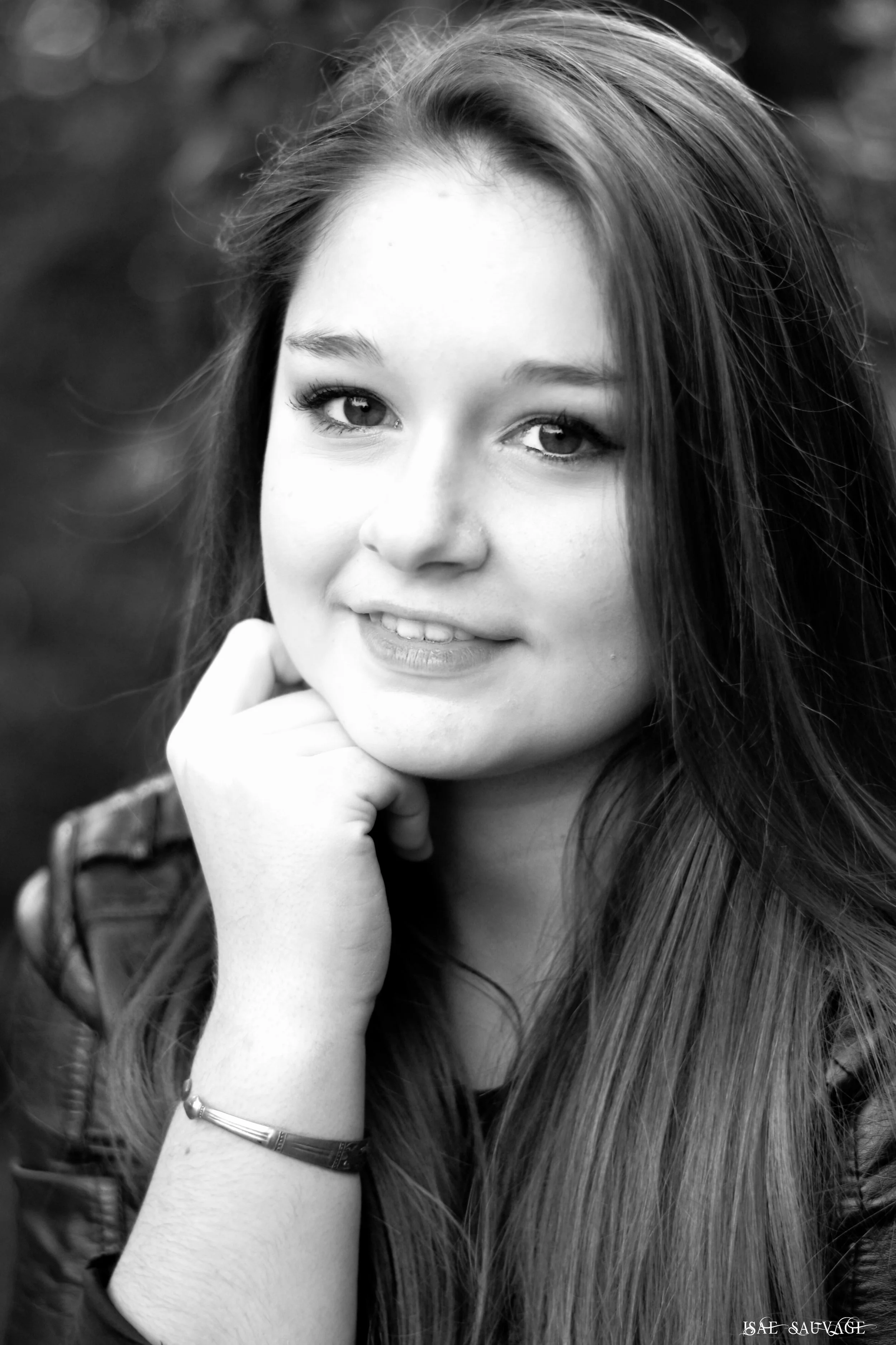 Portrait en noir et blanc d'une jeune femme avec des longs cheveux, qui sourit légèrement et repose sa main sous son menton. Elle porte un bracelet.