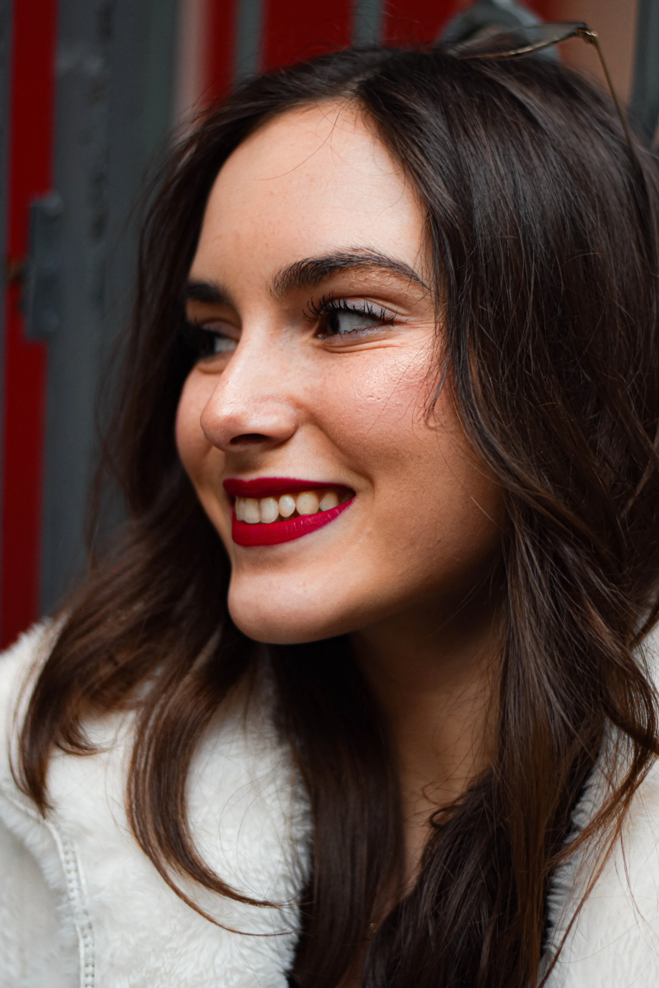 Une jeune femme souriante avec des cheveux bruns longs, portant un rouge à lèvres rouge vif et une veste blanche, regarde légèrement vers la droite.