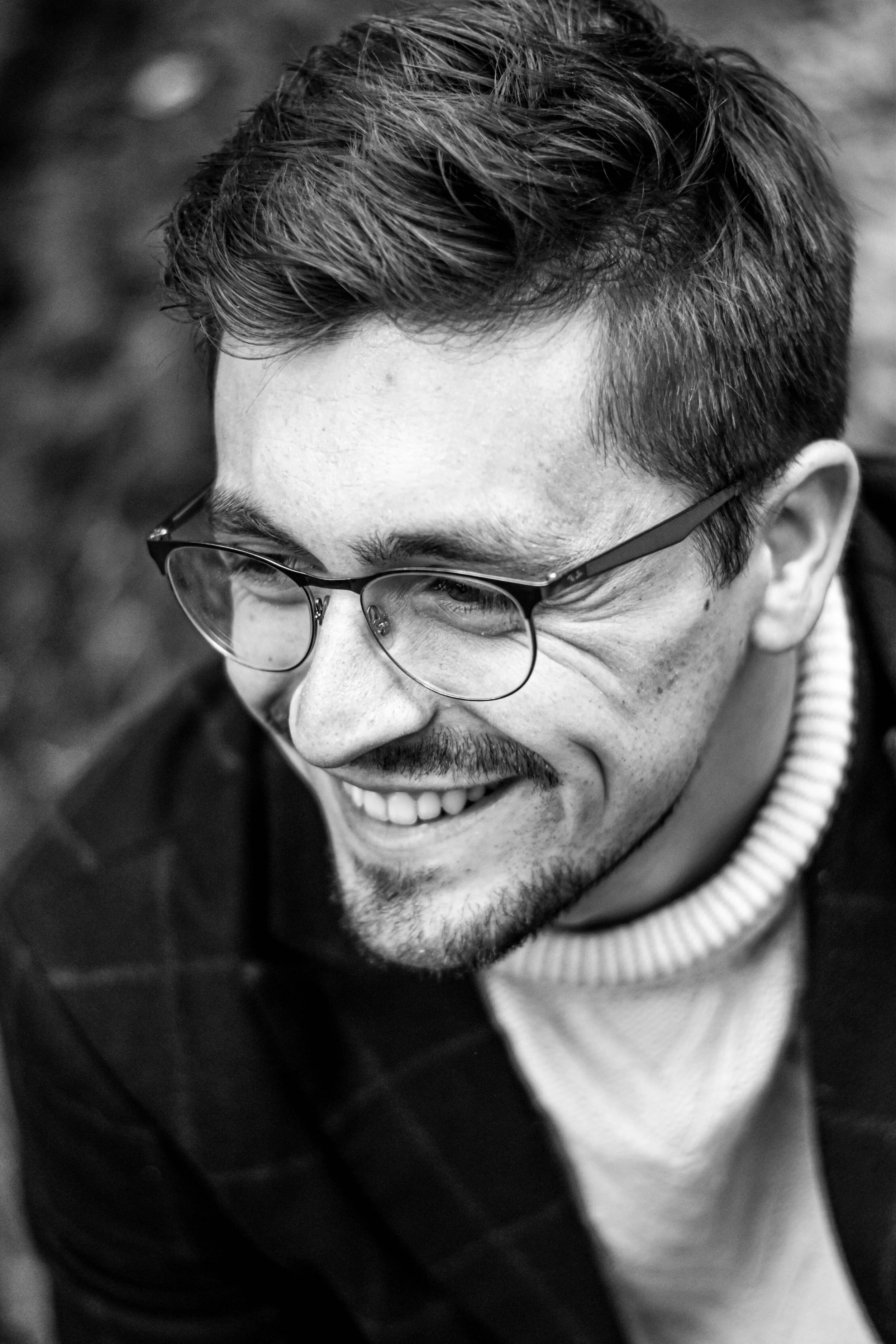Un homme souriant avec des lunettes, court cheveux foncés et barbe, en très proche vue, en noir et blanc.