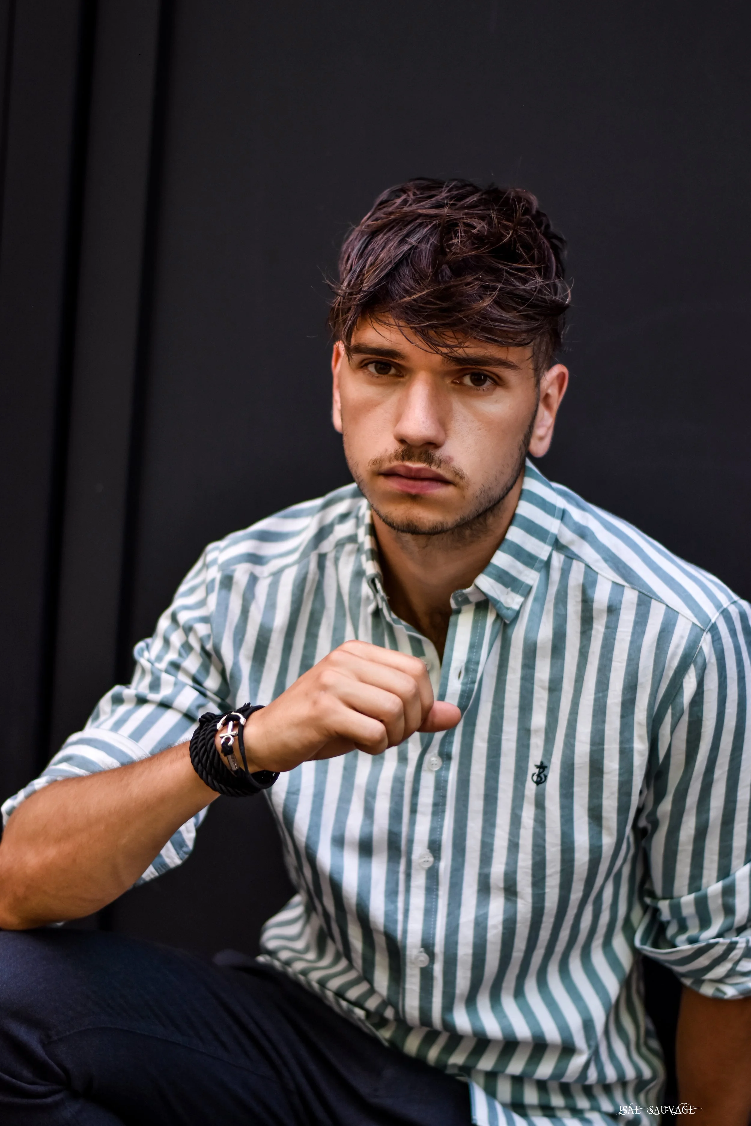 Un jeune homme assis contre un fond noir, portant une chemise rayée vert et blanc, regarde l'objectif avec une expression sérieuse. Il porte un bracelet noir enroulé au poignet gauche et a les cheveux bruns coiffés en désordre.