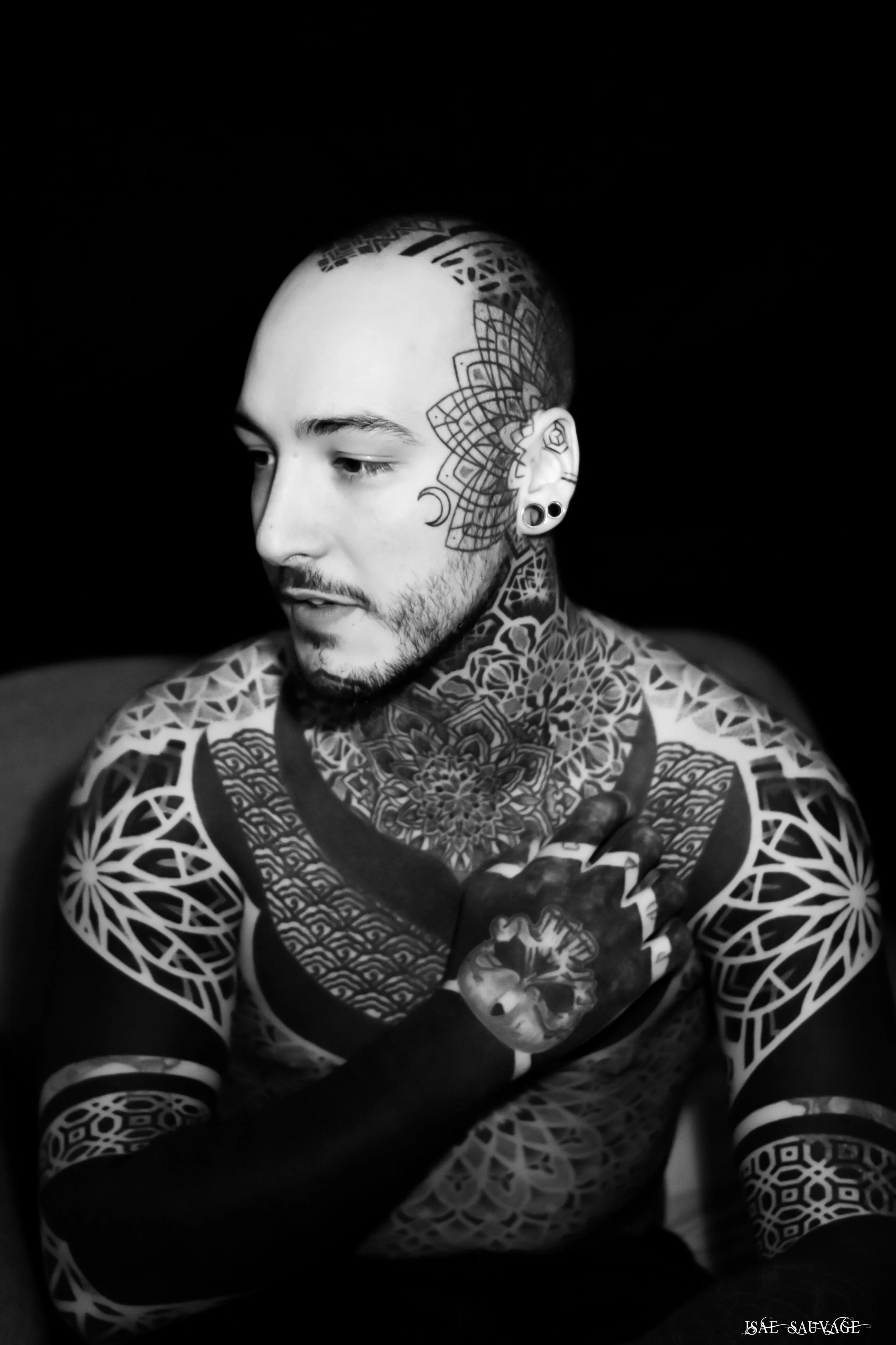 Un homme tatoué avec des motifs géométriques, un visage partiellement vu, portant des piercings à l'oreille, regardant vers la gauche, en noir et blanc.