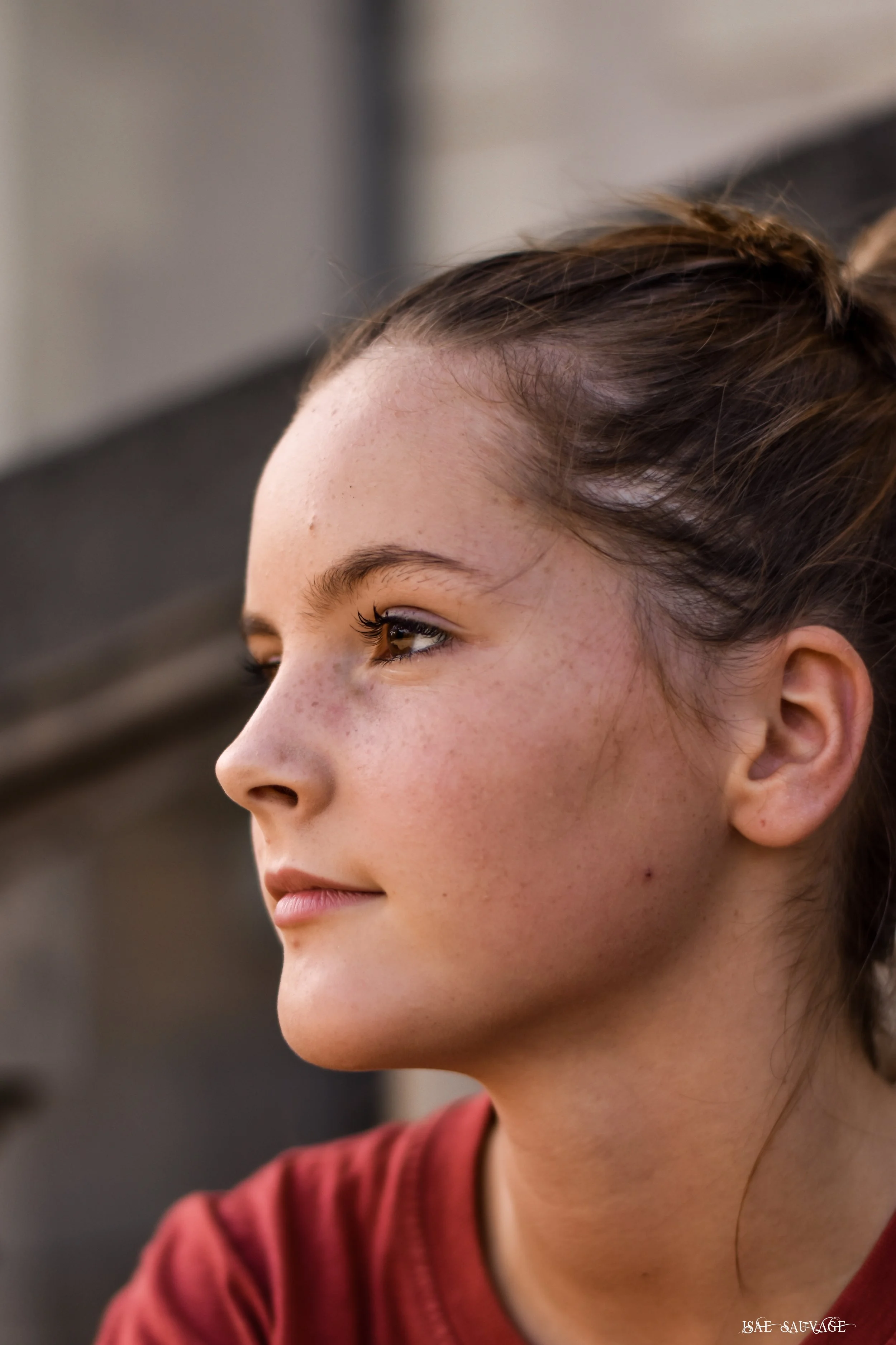 Profil d'une jeune femme aux cheveux en chignon et peau claire, portant un haut rouge, regardant légèrement vers la droite, avec un fond flou.