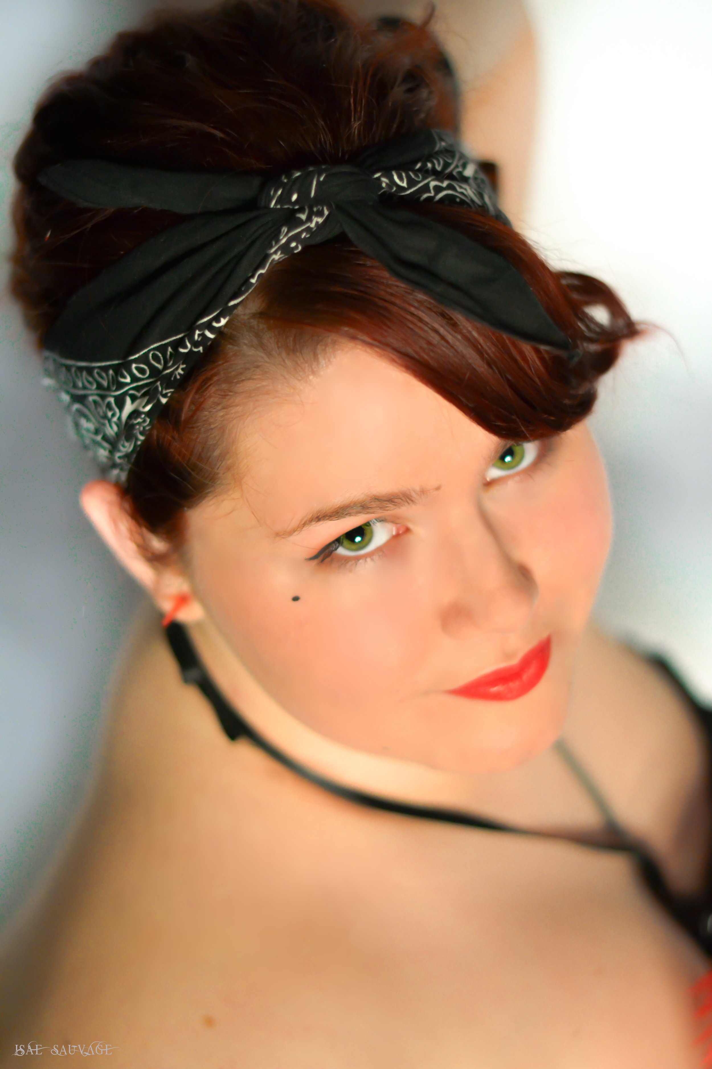 Une jeune femme avec des cheveux roux sombre, portant un bandana noir avec des motifs blancs, voit de près. Elle porte aussi un maquillage rouge à lèvres et un collier noir, avec une peau claire et un petit point noir sous l'œil.