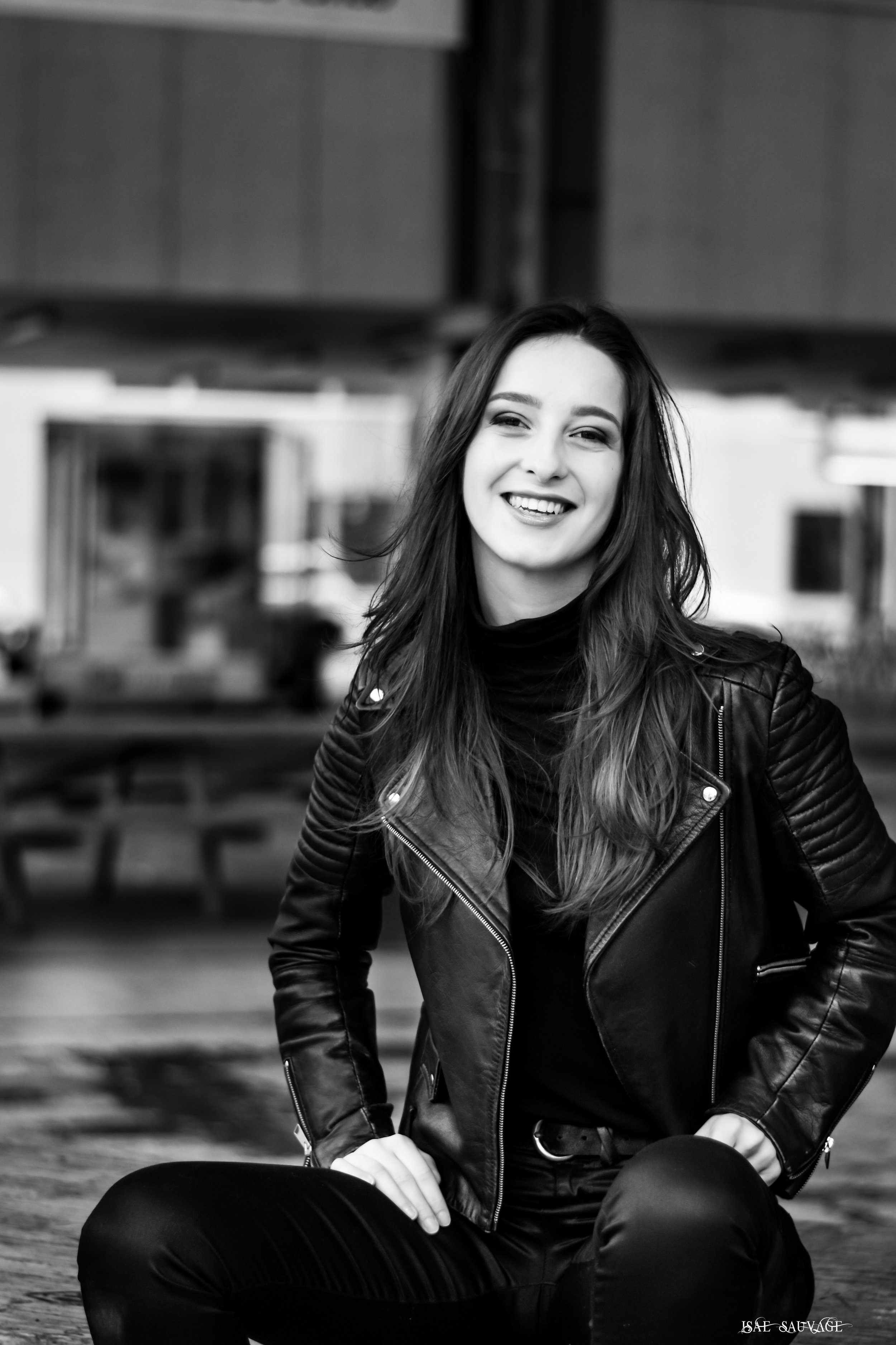 Une femme souriante avec des cheveux longs, portant un blouson en cuir noir, assise en extérieur, en noir et blanc.
