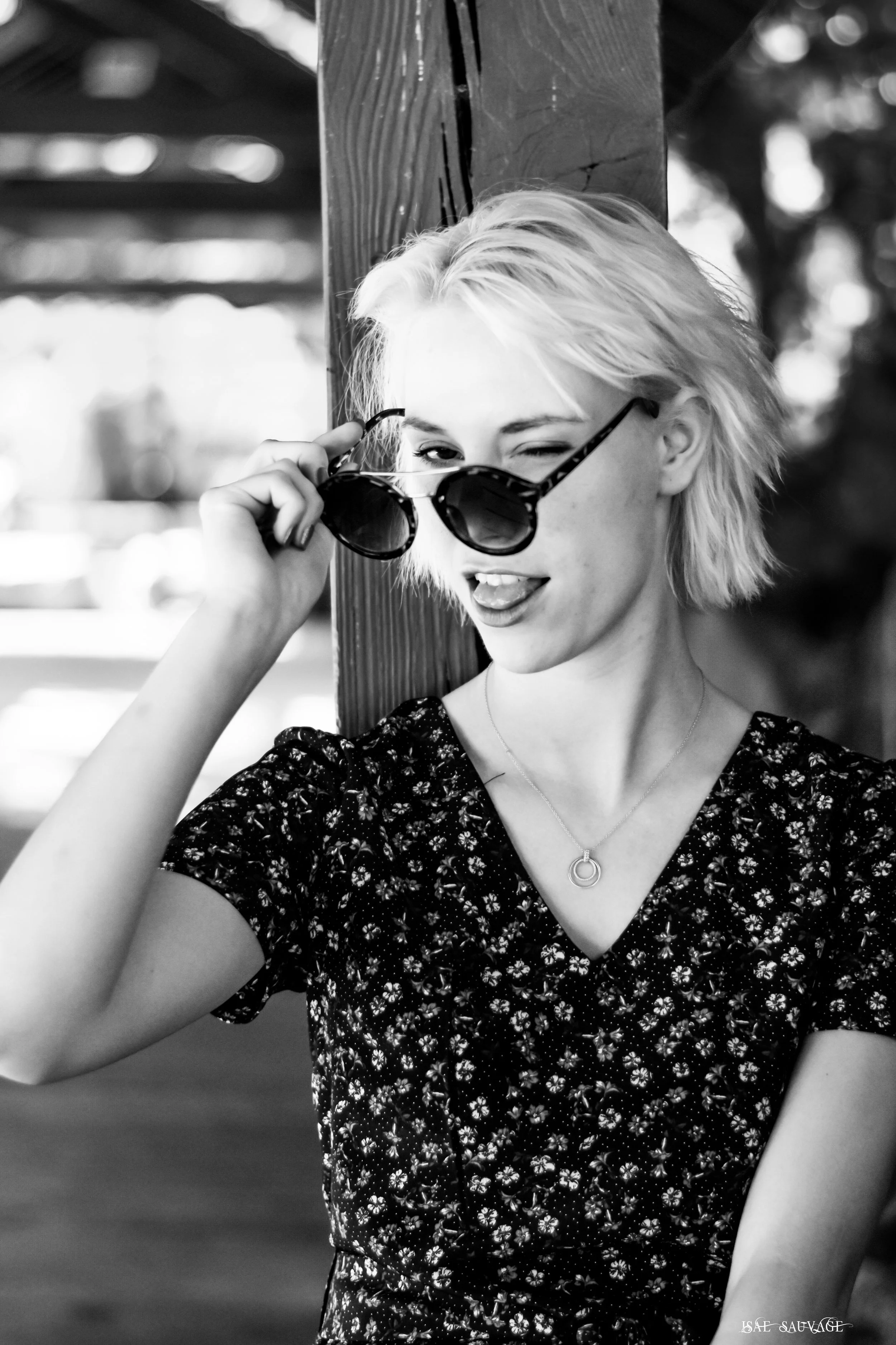 Portrait d'une jeune femme portant des lunettes de soleil, avec une expression souriante et une langue sortie, cheveux courts et clairsemés, vêtue d'une robe à motifs floraux, en noir et blanc.