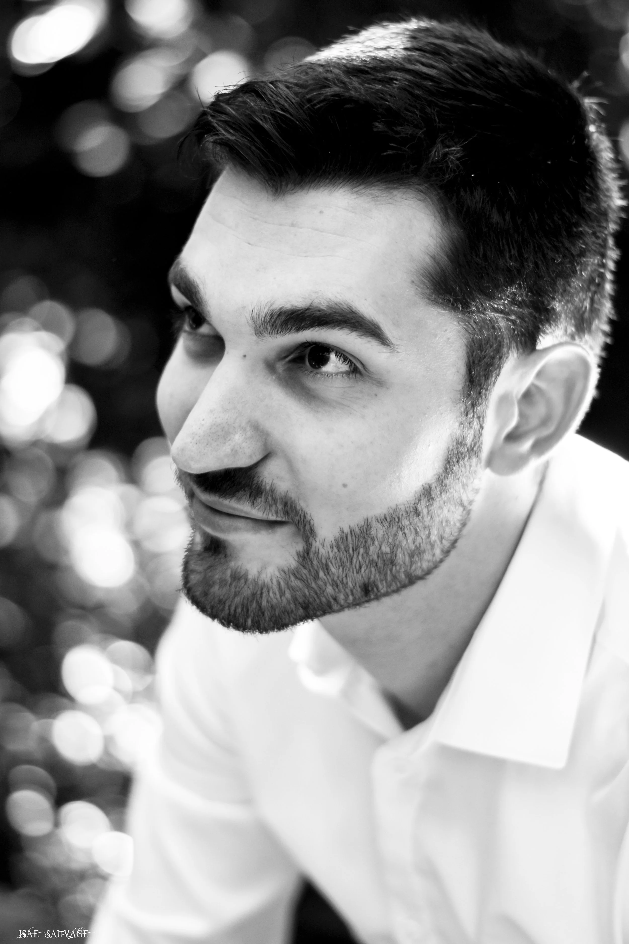 Portrait en noir et blanc d'un jeune homme avec barbe, regardant vers la gauche, avec un fond flou de lumières brillantes.