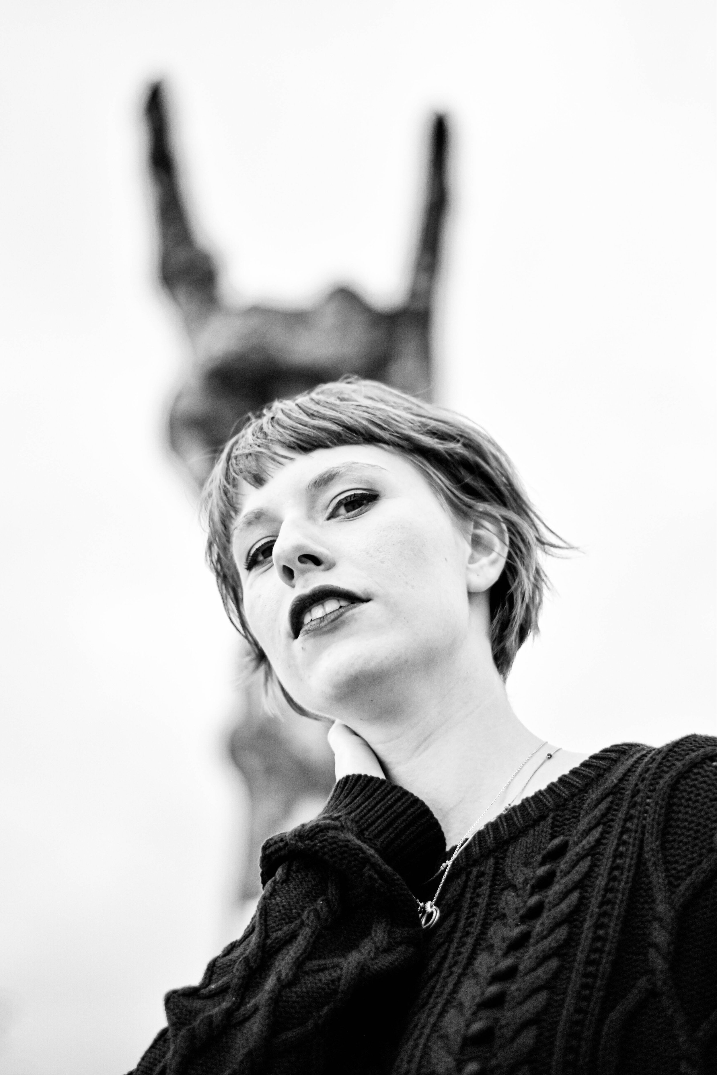 Une jeune femme avec des cheveux courts et un sourire, porte une veste en tricot épais, avec un fond flou montrant une sculpture du parc du Hellfest