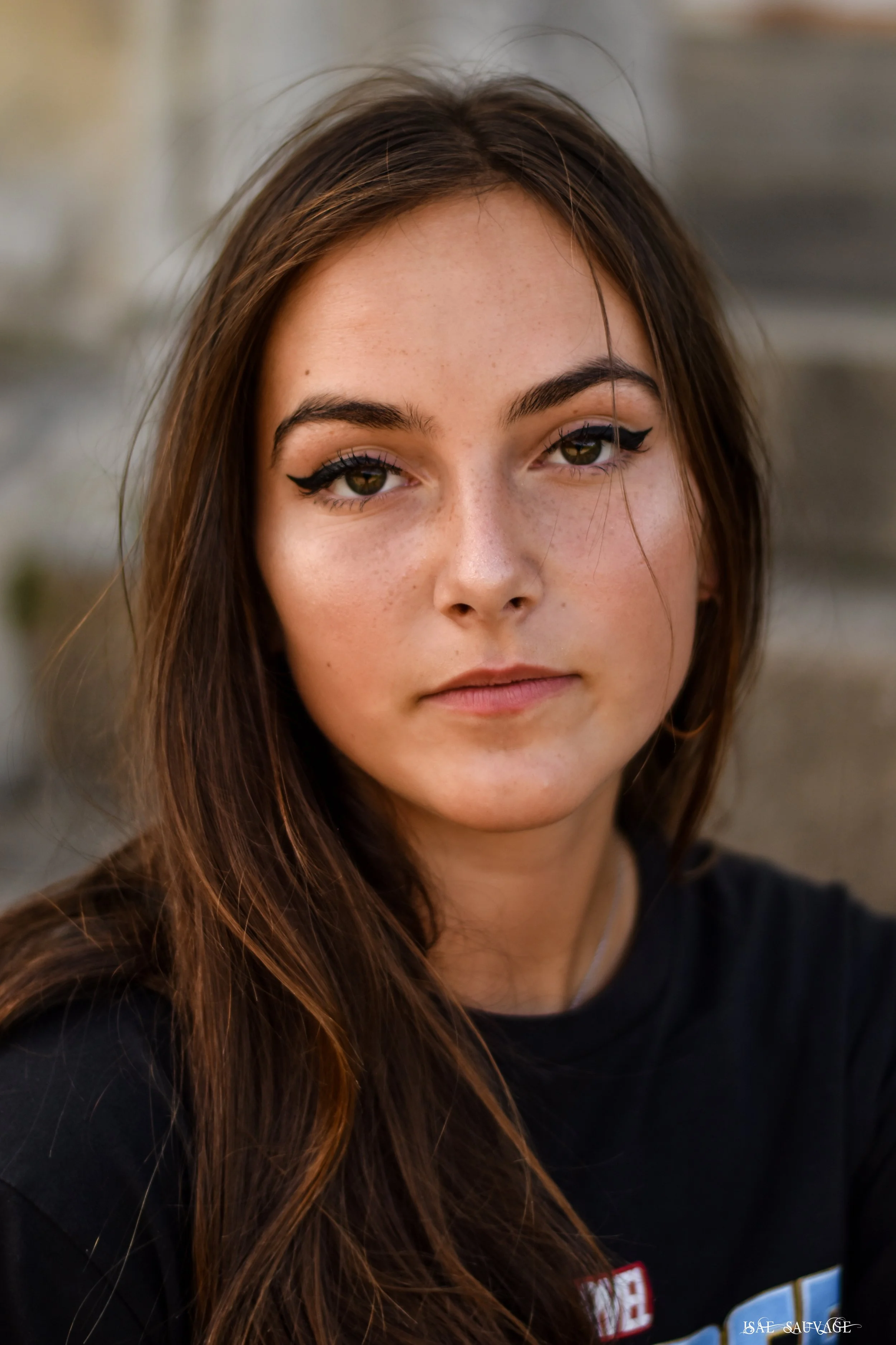 Portrait d'une jeune femme avec des cheveux longs, porte un maquillage avec un eyeliner noir et a des taches de rousseur, portait un haut noir.