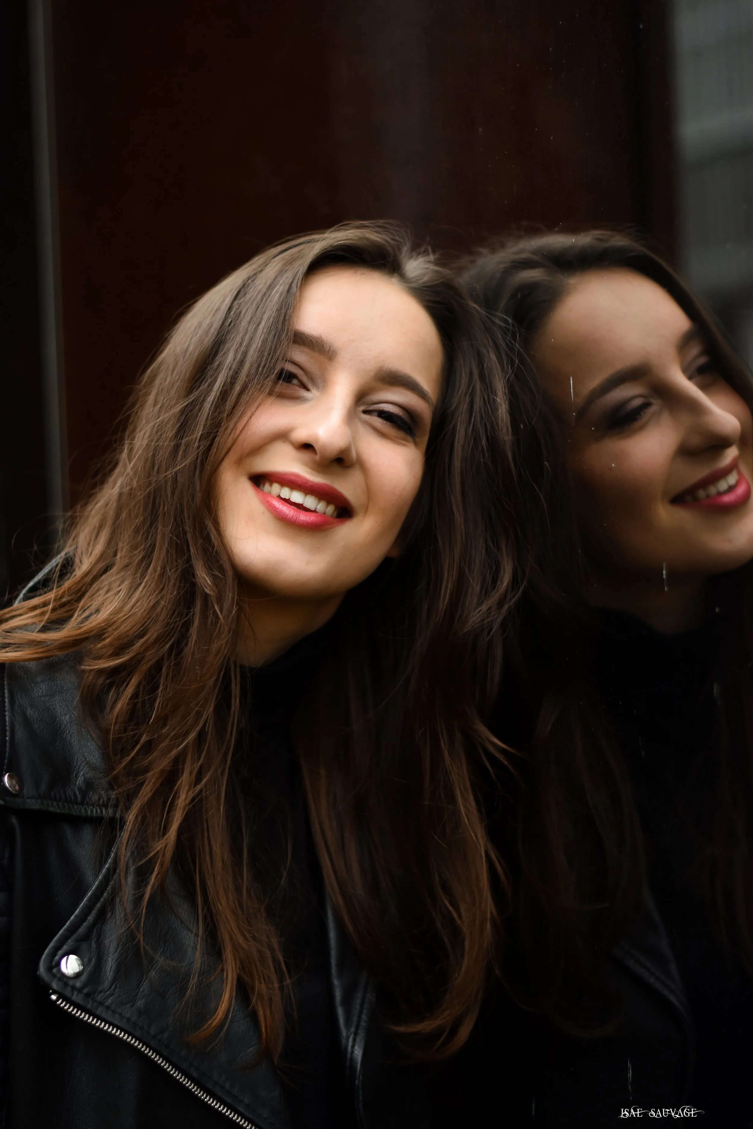 une jeune femme avec long cheveux bruns souriante, regardant son reflet dans une vitre de batiment, vestimentaire décontracté. 