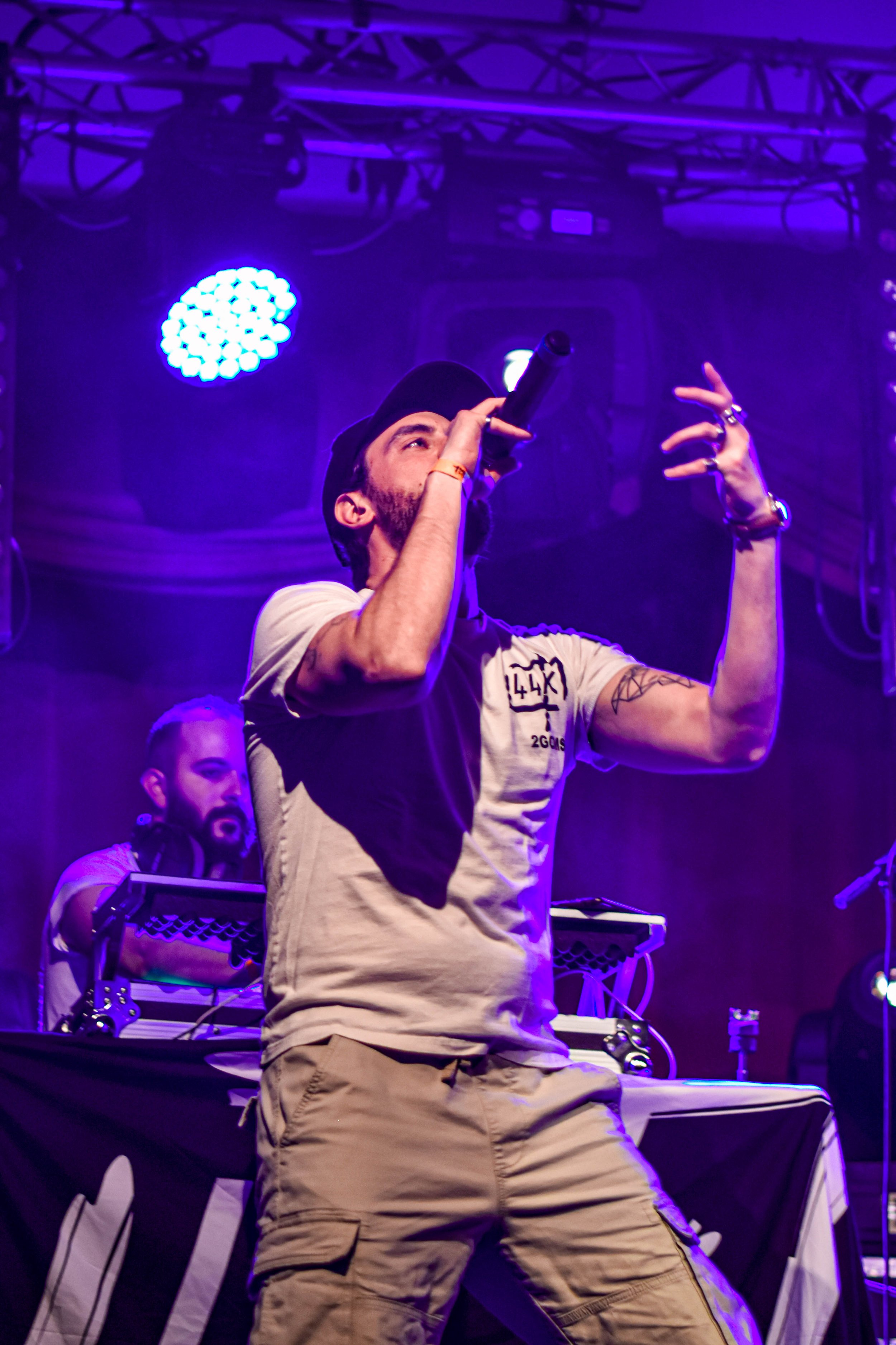 Un homme en t-shirt beige, portant un chapeau noir, chante dans un microphone lors d'un concert, avec des lumières violettes en arrière-plan.