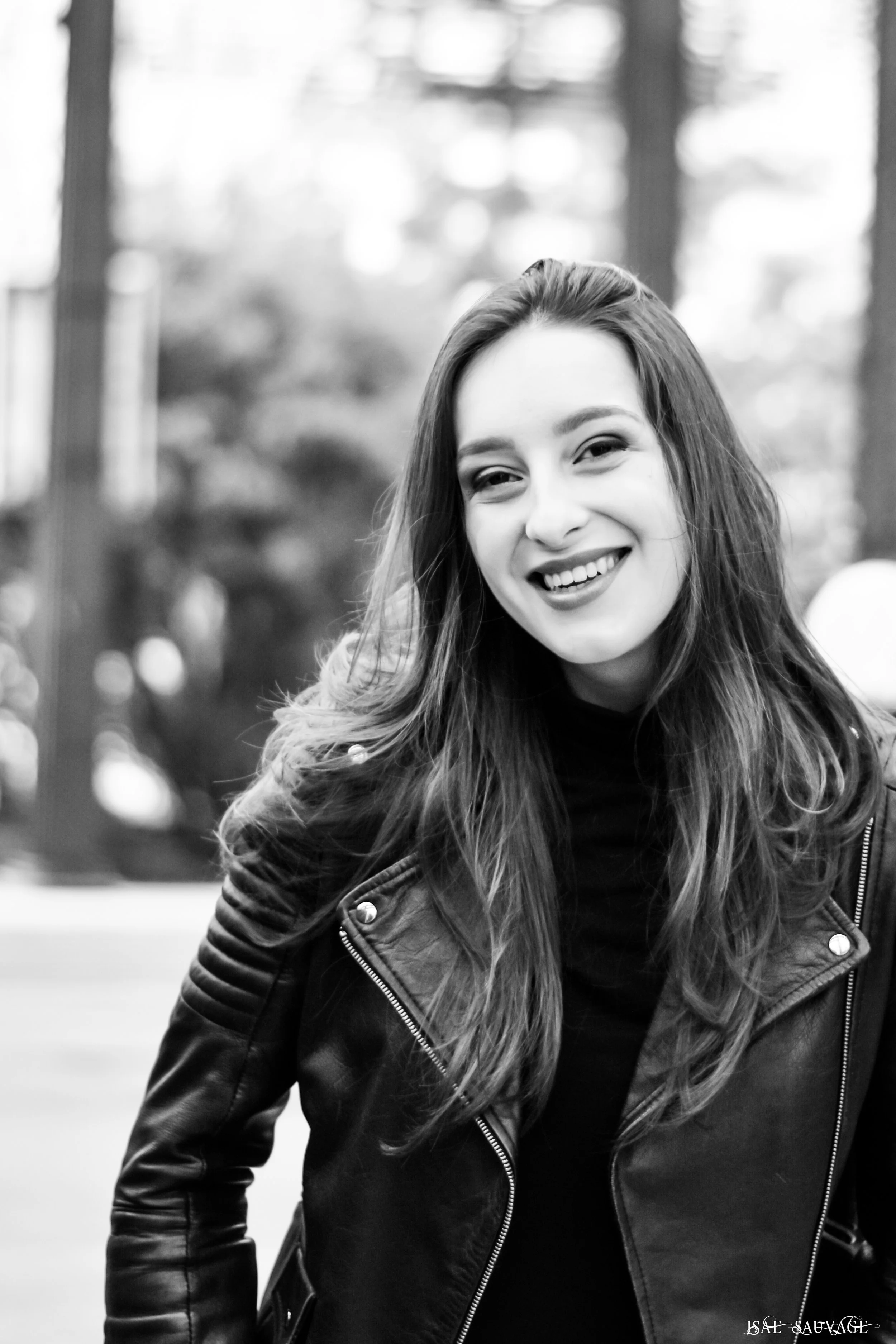 Portrait d'une jeune femme souriante avec des cheveux longs et bouclés, portant une veste en cuir, dans un environnement extérieur flou.