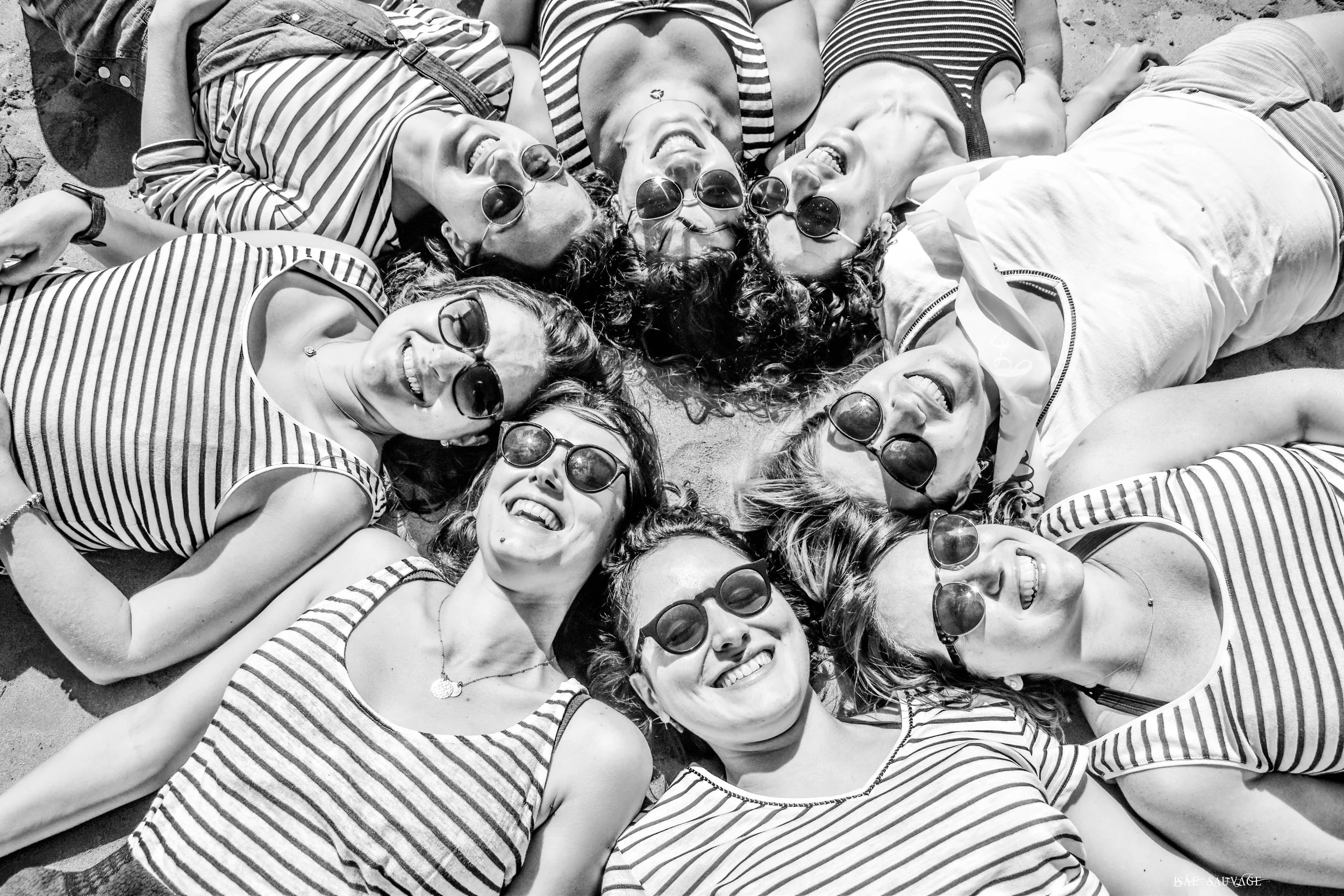 Groupe de personnes allongées sur le sable souriantes, portant des vêtements à rayures et des lunettes de soleil.