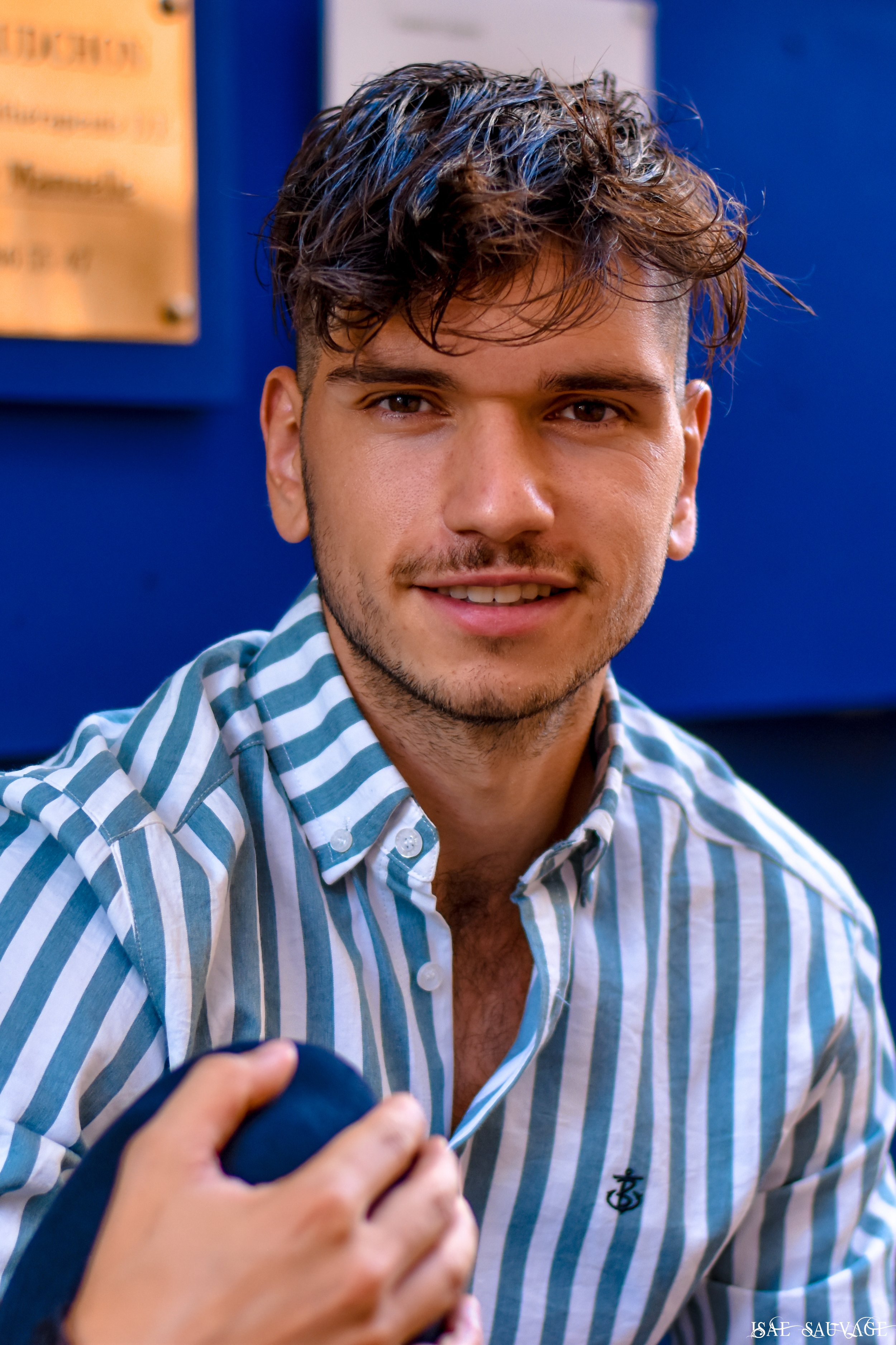 Un jeune homme avec des cheveux bruns et ondulés, portant une chemise à rayures vertes, blanches et bleues, sourit légèrement en concentrant son regard. 