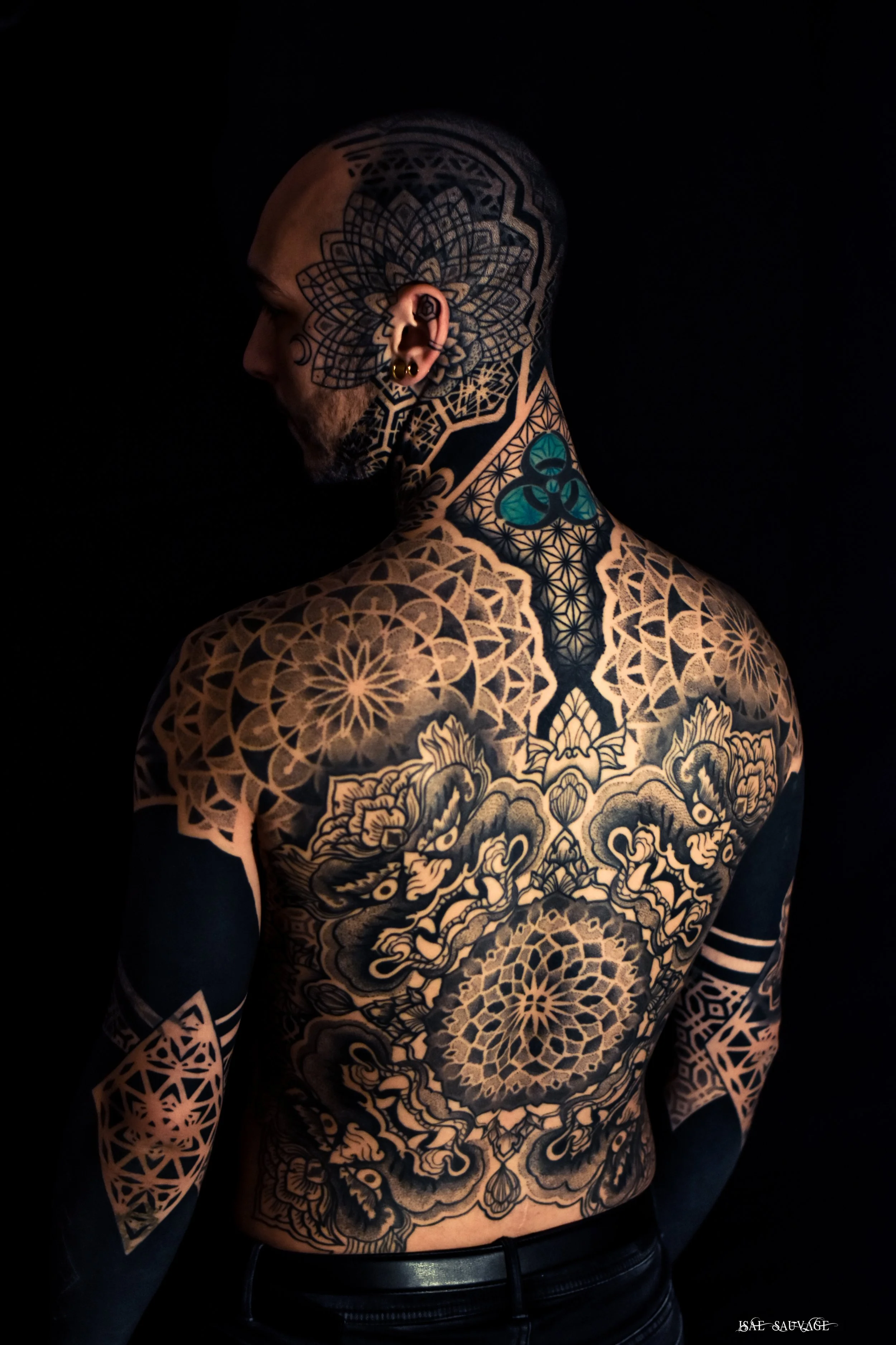 Un homme sans cheveux, vu de profil, avec de nombreux tatouages noirs et bleus couvrant sa tête, son cou, ses bras et son dos, comprenant des motifs géométriques et floraux, dans un fond noir.