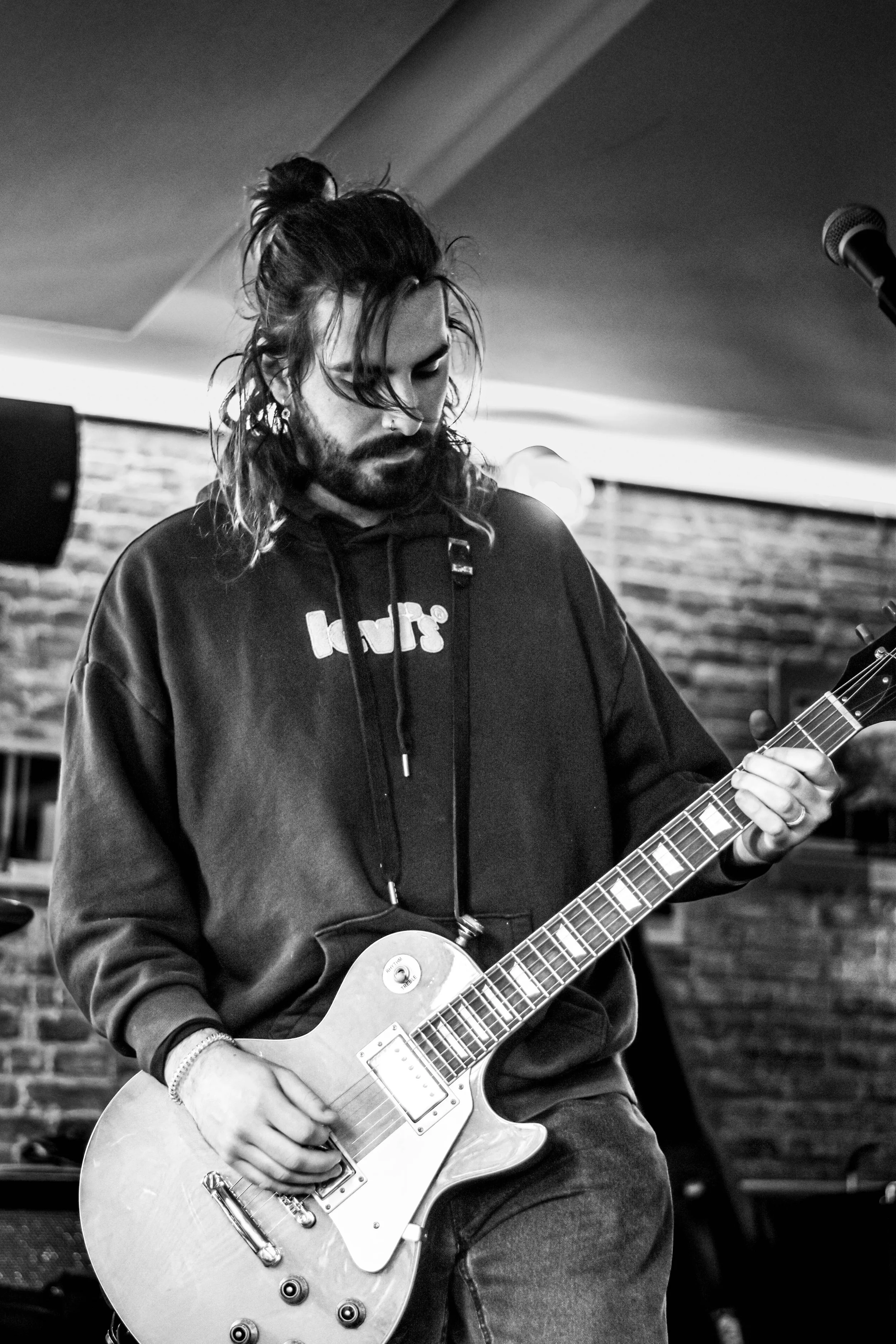 Un homme avec une barbe et des cheveux longs attachés joue de la guitare électrique en noir et blanc dans un intérieur avec un mur en briques.