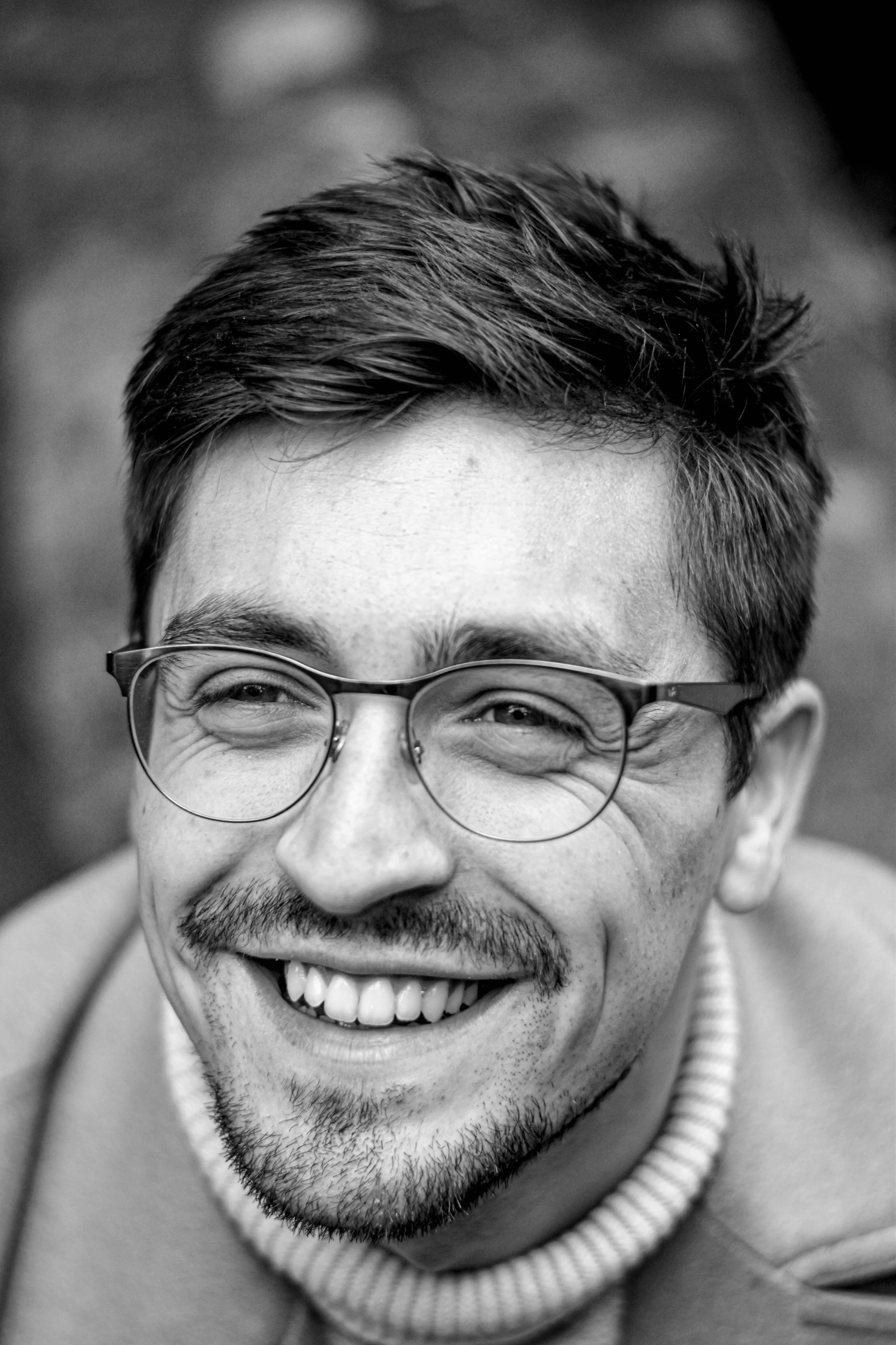 Sourire d'un homme portant des lunettes, sans barbe, avec une coiffure courte et une barbe légère, en noir et blanc.