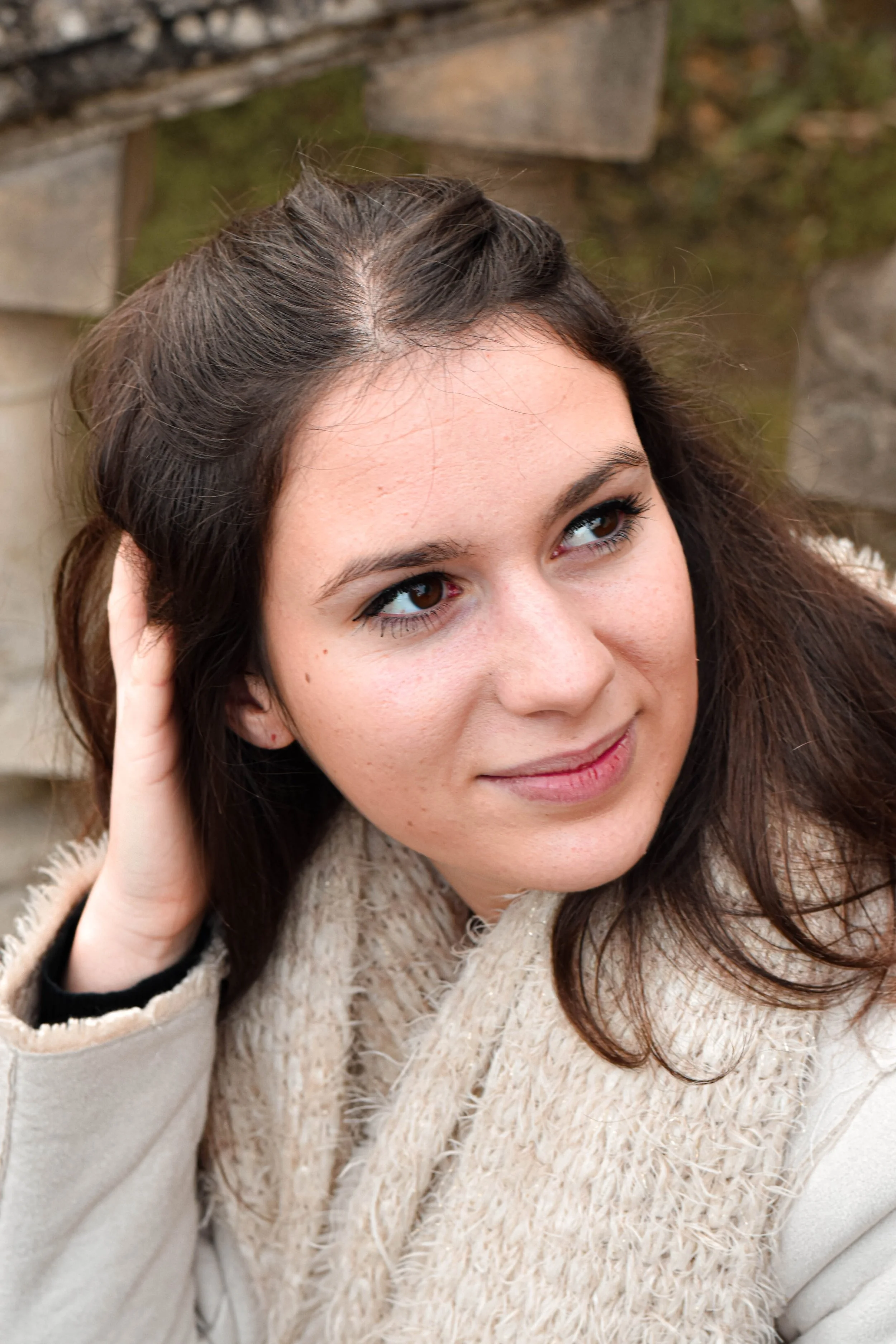Une femme aux cheveux bruns, portant un pull beige avec une écharpe en laine, face à l'objectif, dans un décor en pierre et bois.