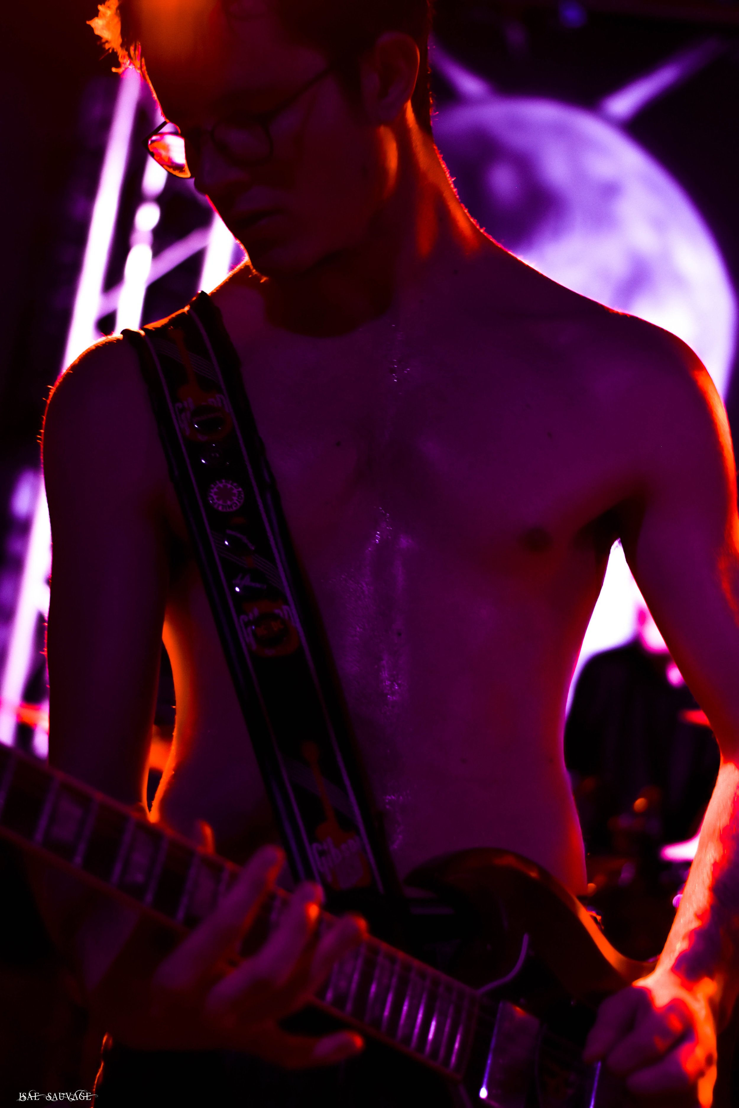 Une personne torse nue, portant des lunettes, jouant de la guitare lors d'un concert dans une ambiance colorée et sombre