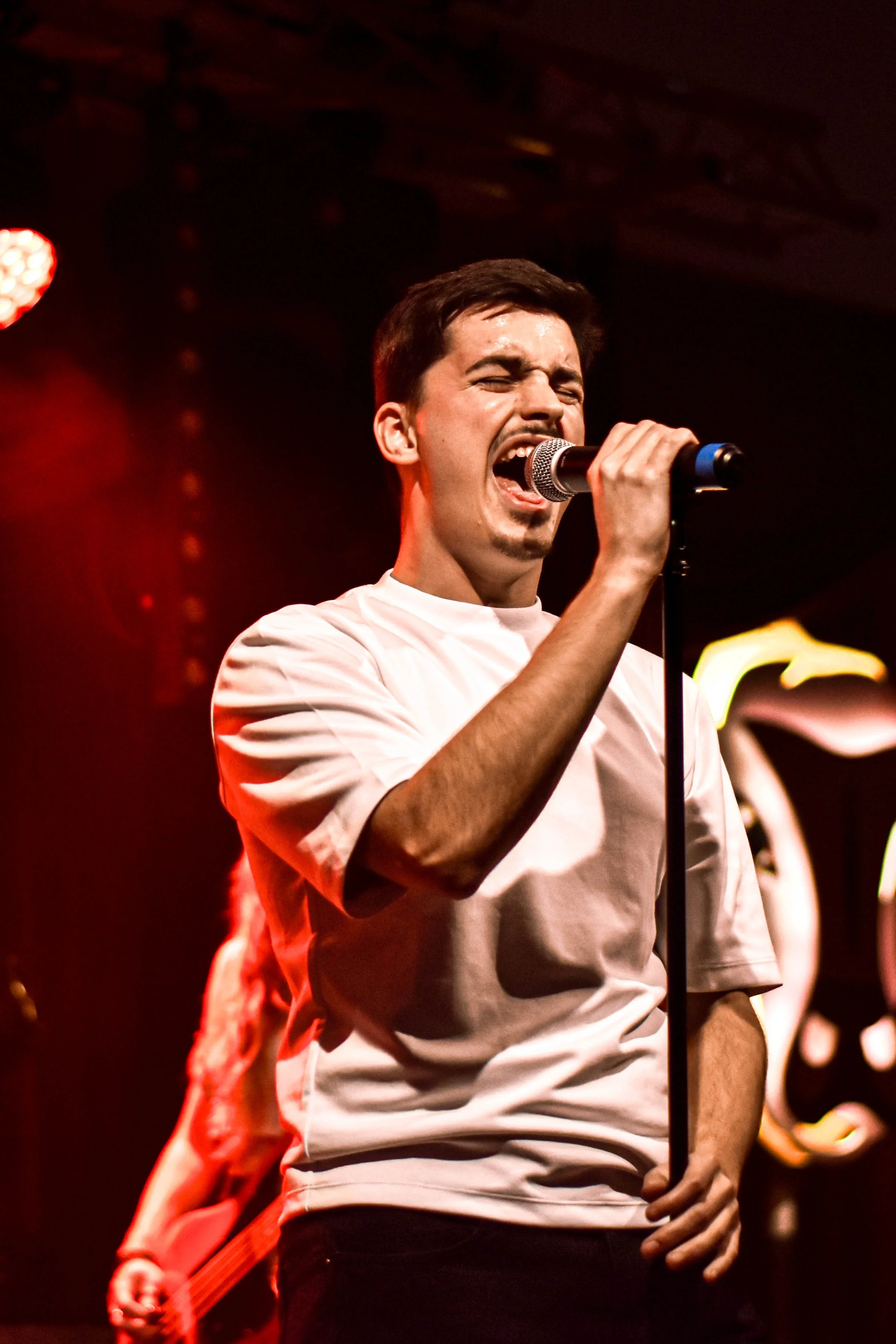 Un homme chante dans un micro lors d'un concert, avec un fond sombre et des lumières rouges et jaunes. Il porte un t-shirt blanc et a les yeux fermés, montrant une forte émotion.