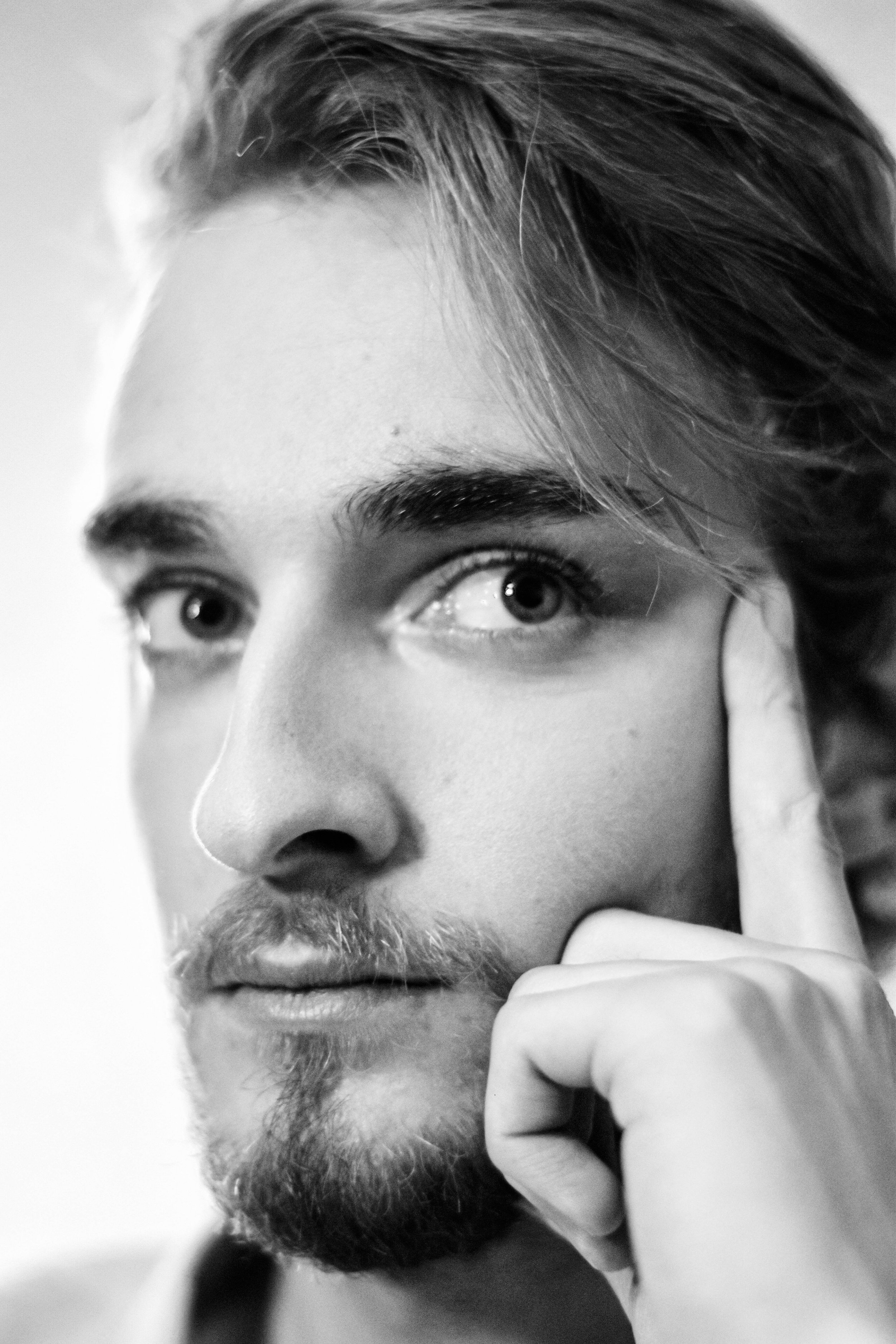 Un portrait en noir et blanc d'un jeune homme avec une barbe et une moustache, regardant vers le haut, tenant son menton avec sa main, avec ses cheveux au style décontracté.