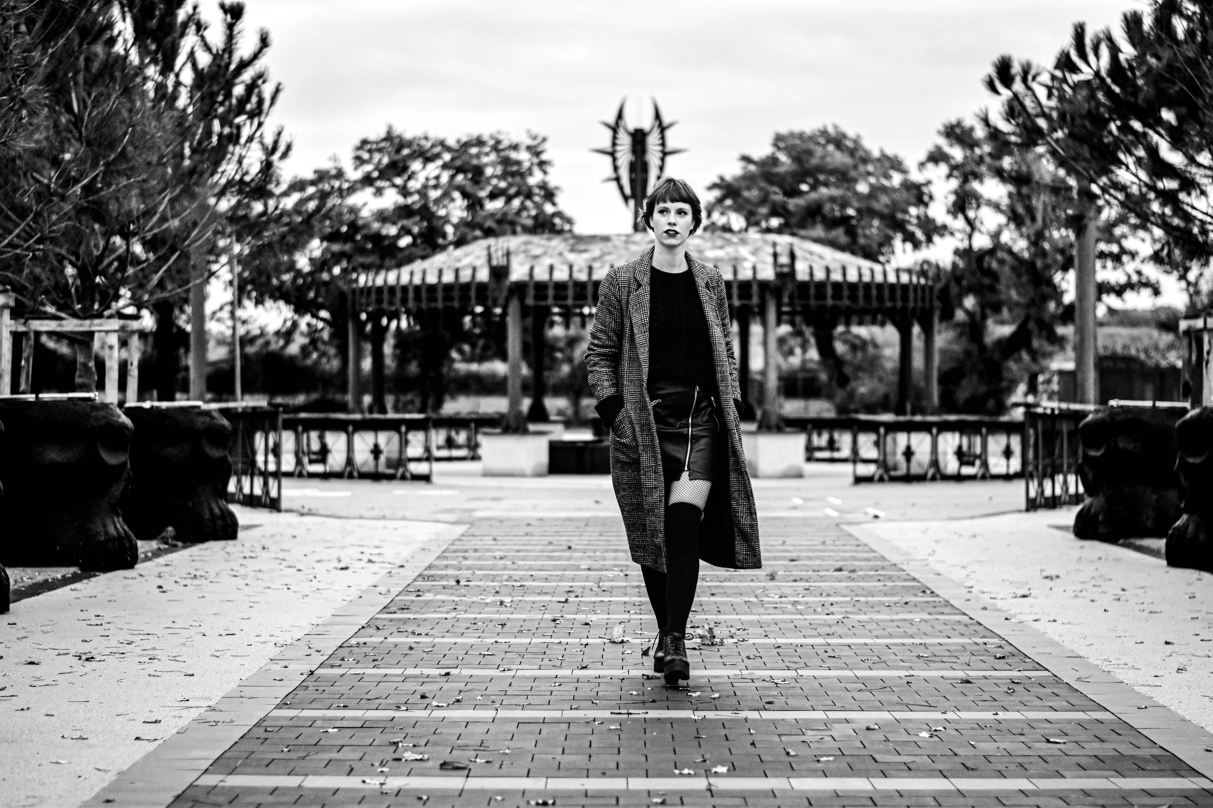 Une femme marche seul sur une allée pavée dans un parc, portant un manteau à carreaux, des bottes noires et une jupe courte, avec des arbres en arrière-plan et une structure en forme de totem ou sculpture derrière elle.