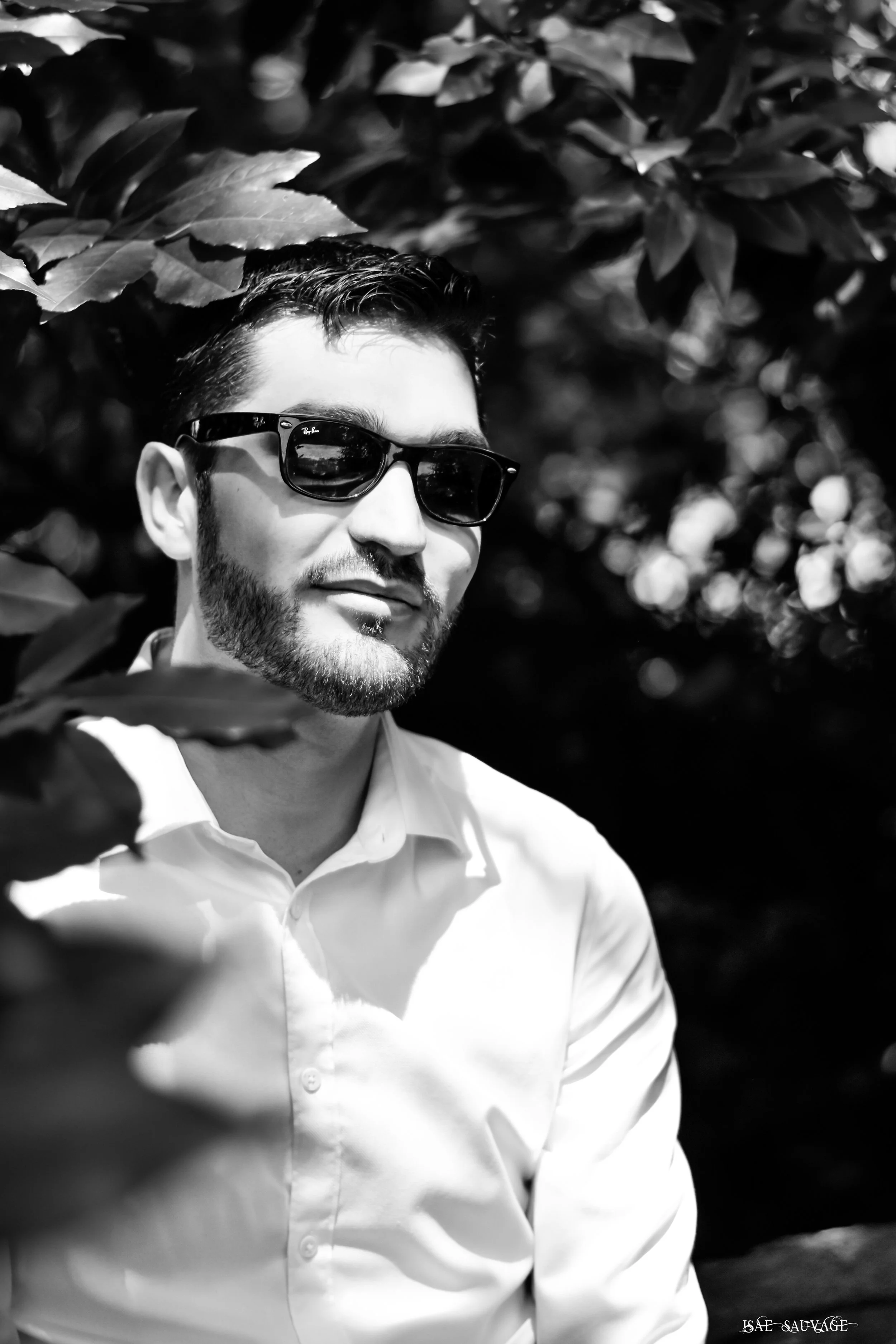 Portrait d'un homme portant des lunettes de soleil Ray-Ban, portant une chemise blanche, dans un environnement avec des feuillages, en noir et blanc.