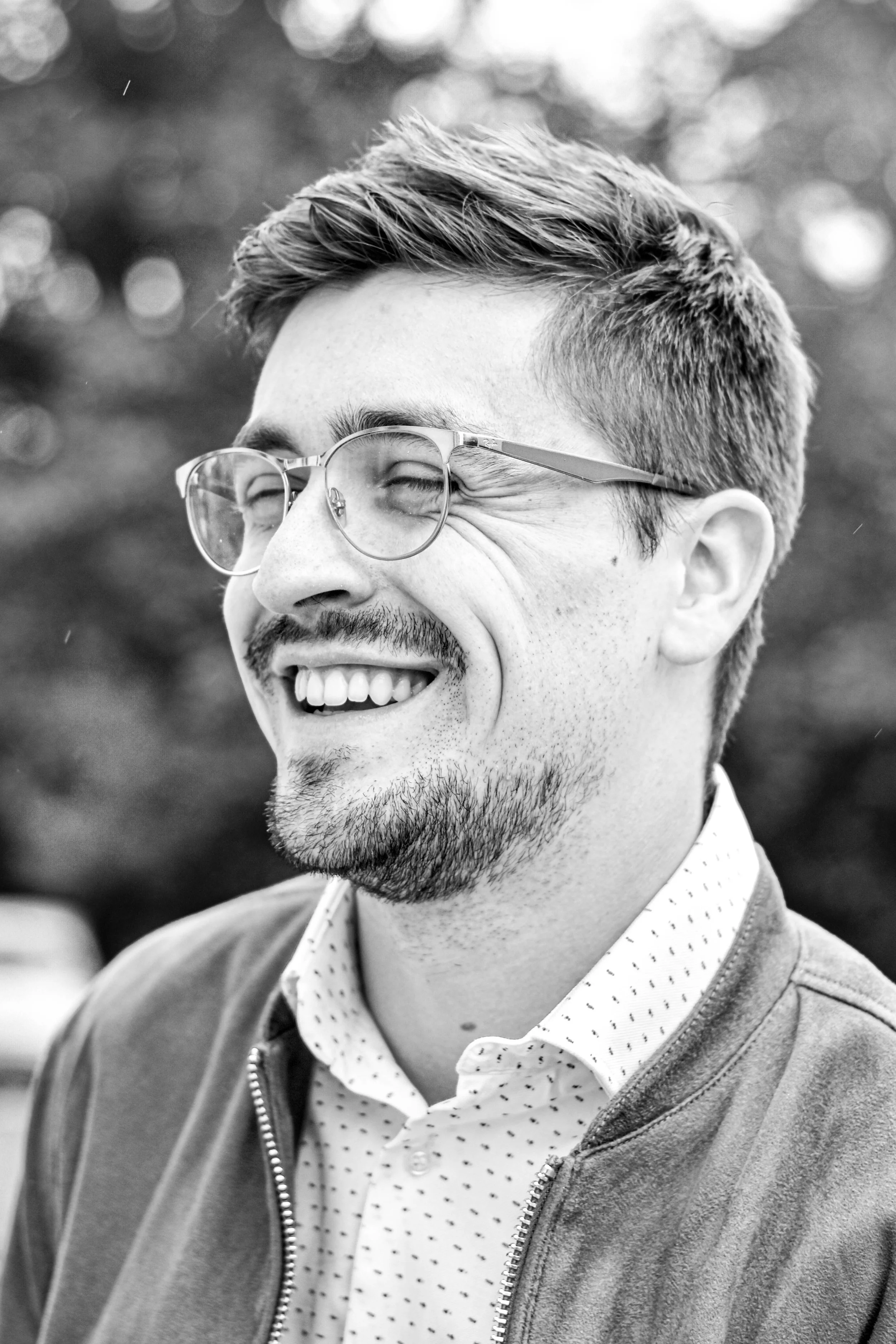 Un homme souriant avec des lunettes, portant une robe à manches longues à motifs, une veste en molleton et une chemise. Il a une barbe légère et des cheveux courts. L'arrière-plan est flou, probablement un extérieur.