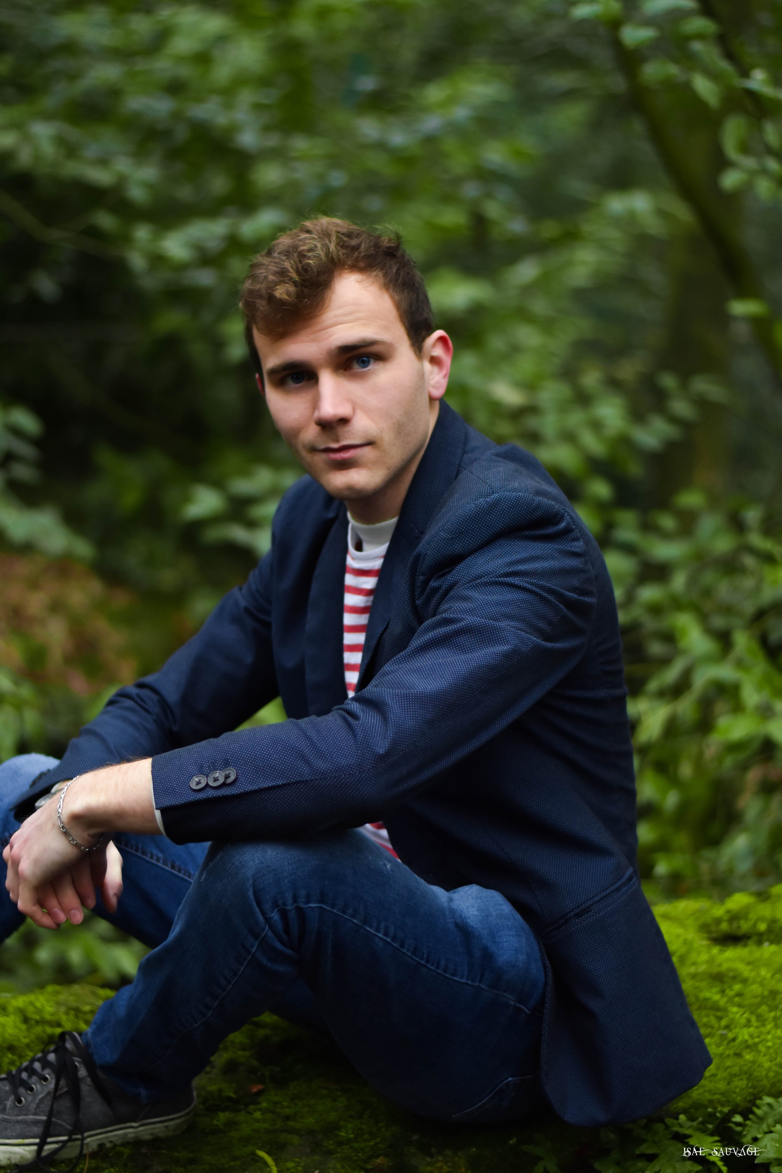Un jeune homme aux cheveux bouclés et aux yeux bleus, portant un blazer bleu, un t-shirt rayé et un jean, assis dans une forêt verte