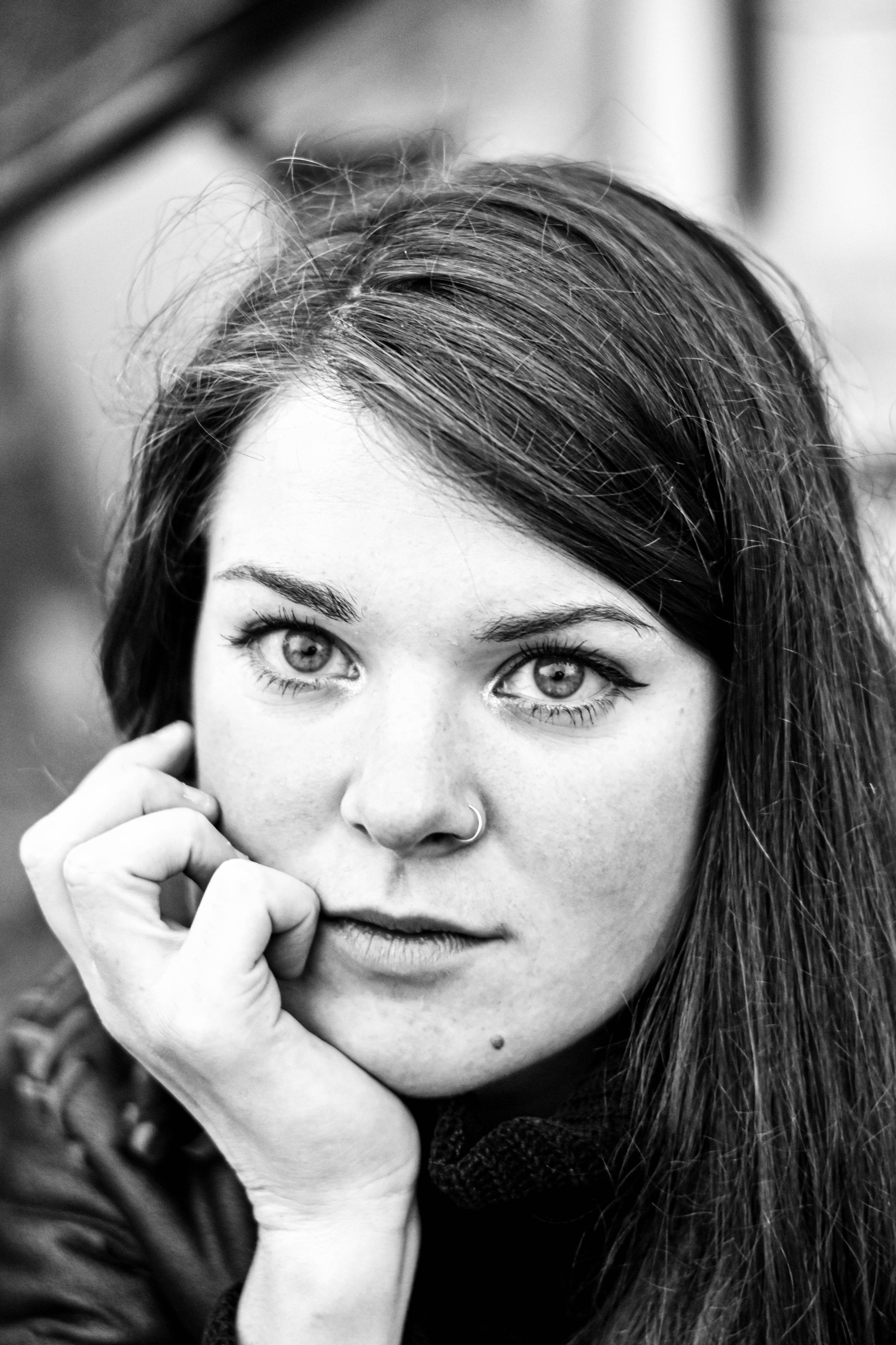 Jeune femme aux yeux bleus, cheveux bruns, portant un piercing au nez, se tenant près d'une barrière, en noir et blanc.