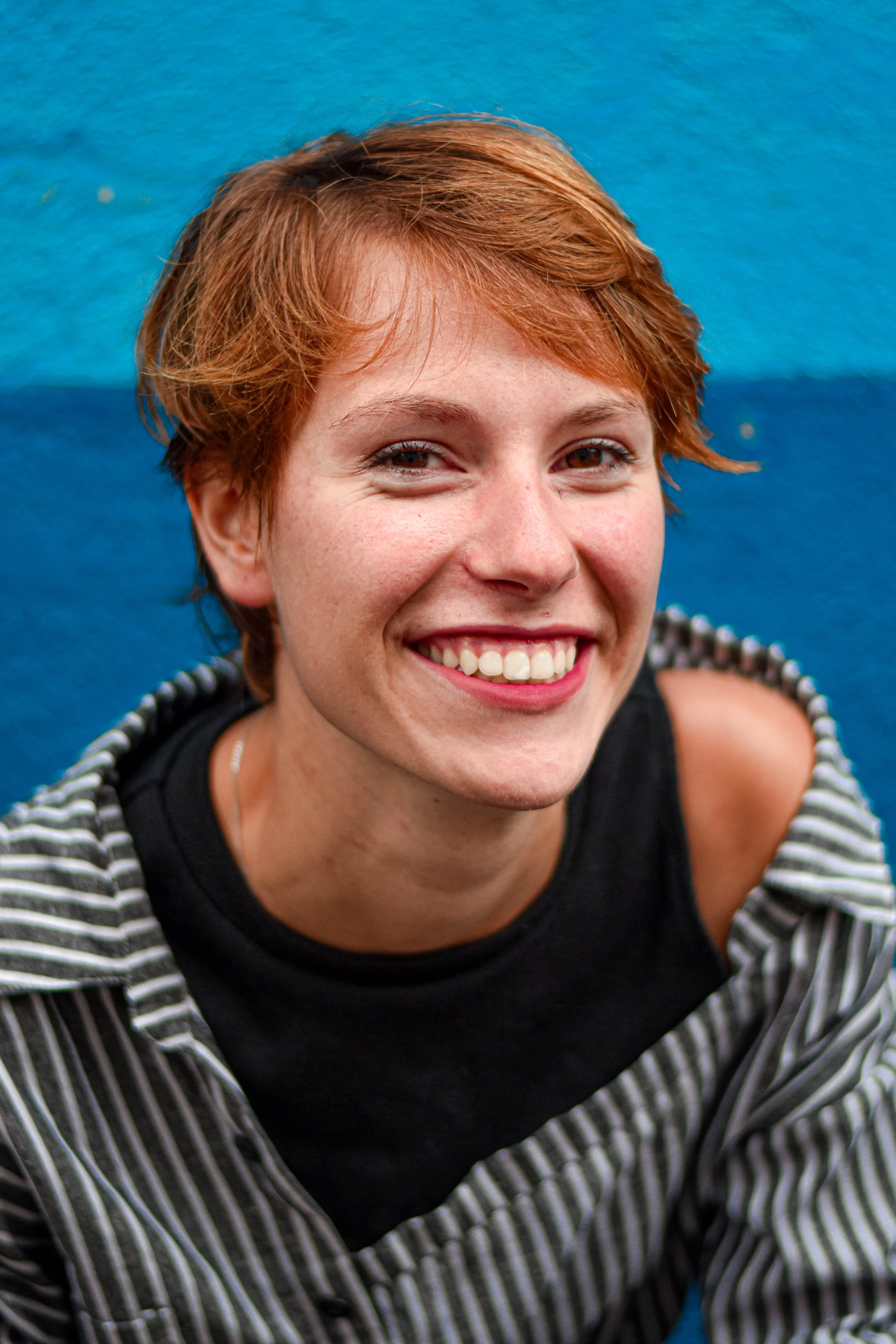 Jeune femme souriante avec cheveux courts et roux, portant un t-shirt noir et une veste à rayures, contre un fond bleu.