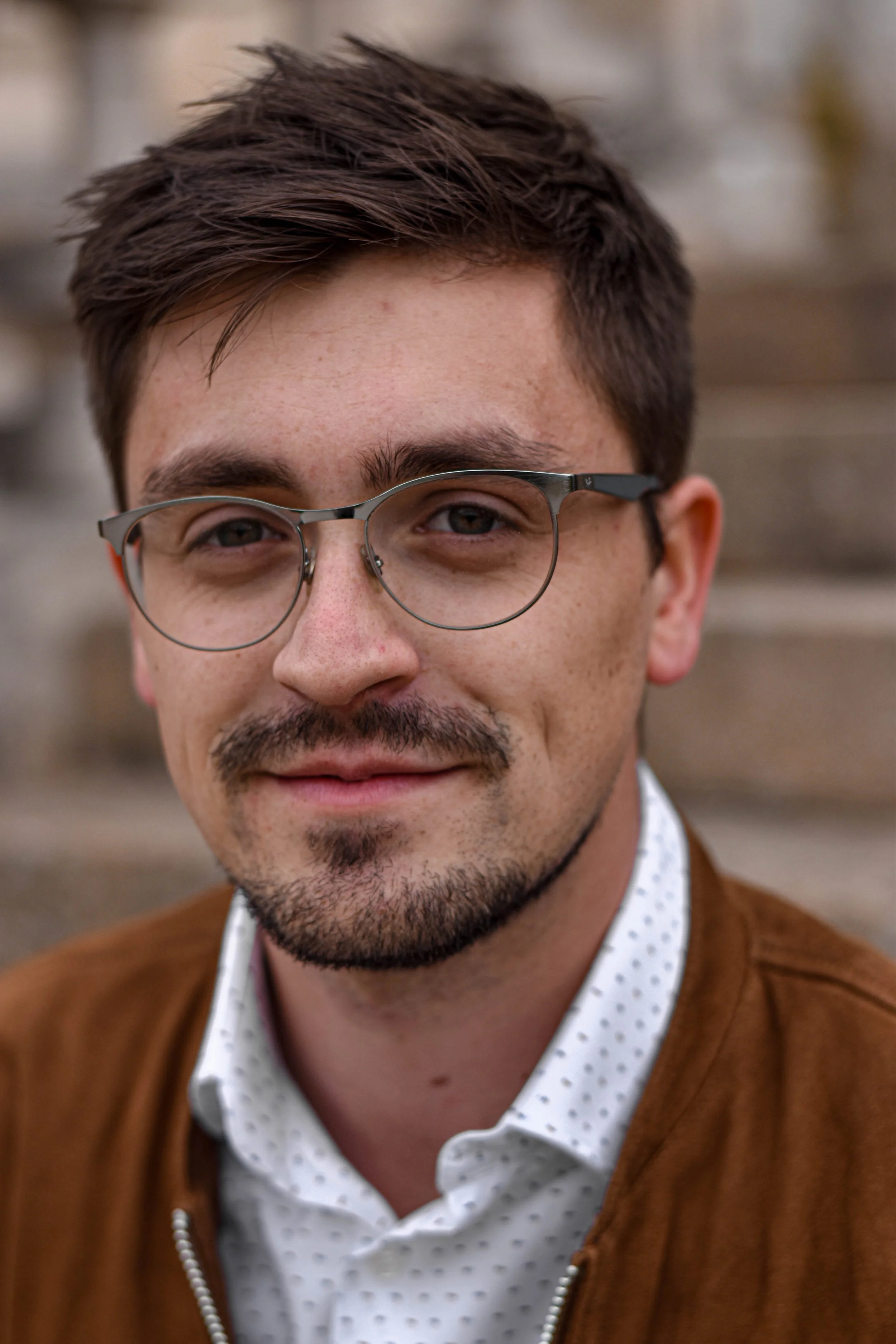 Un homme avec des lunettes, une barbe et une moustache, portant une chemise à pois blancs et noirs sous un vêtement marron, souriant légèrement.